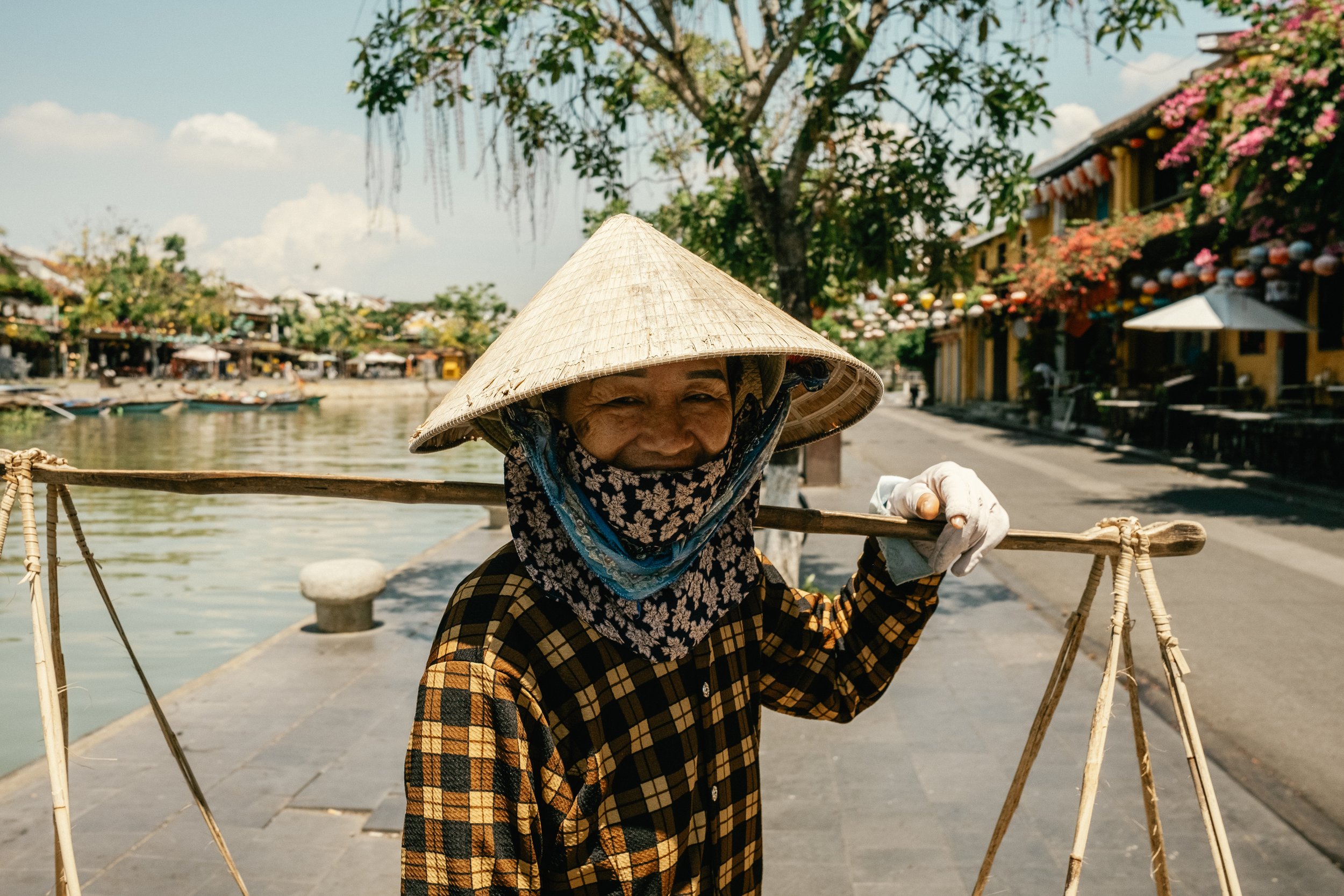 Une femme portant un chapeau conique vietnamien et un foulard couvre son visage, portant un bâton sur ses épaules en bord de rivière dans un village coloré.