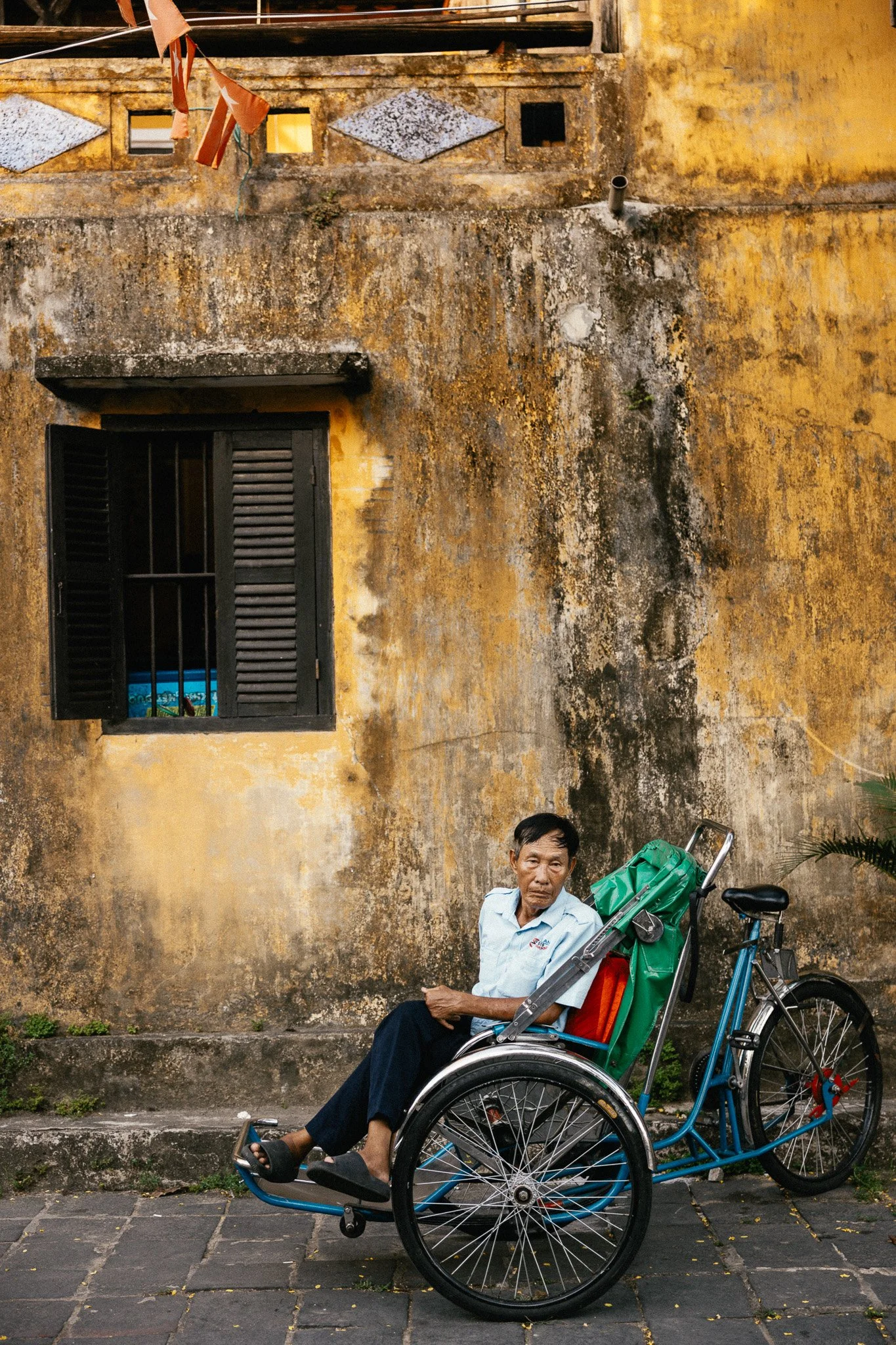 Un homme assis sur un pousse-pousse devant un mur jaune usé avec une fenêtre à volet noir détaché.