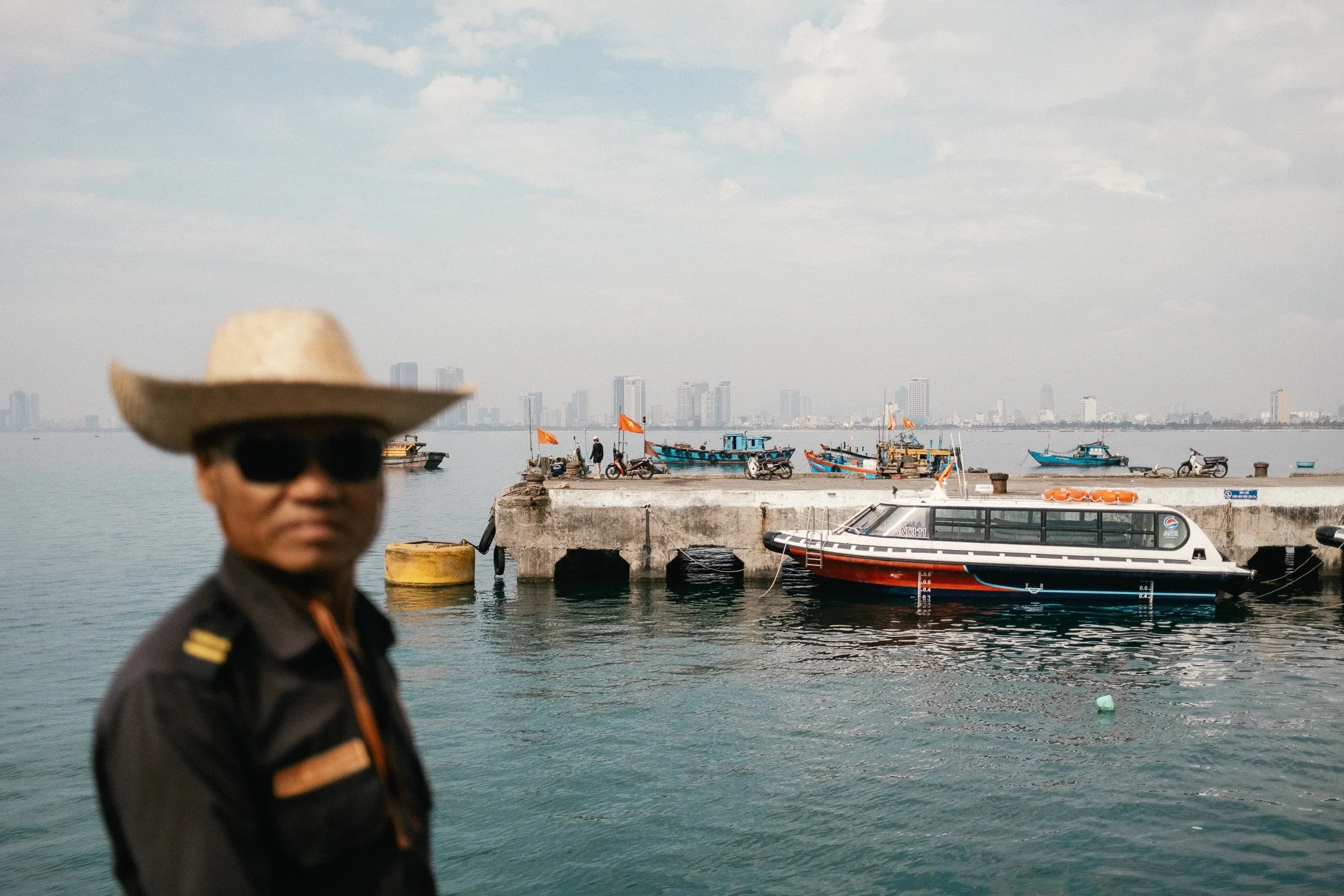 Un homme portant un chapeau de paille et des lunettes de soleil se tient près d'un port avec des bateaux amarrés, une ville en arrière-plan.