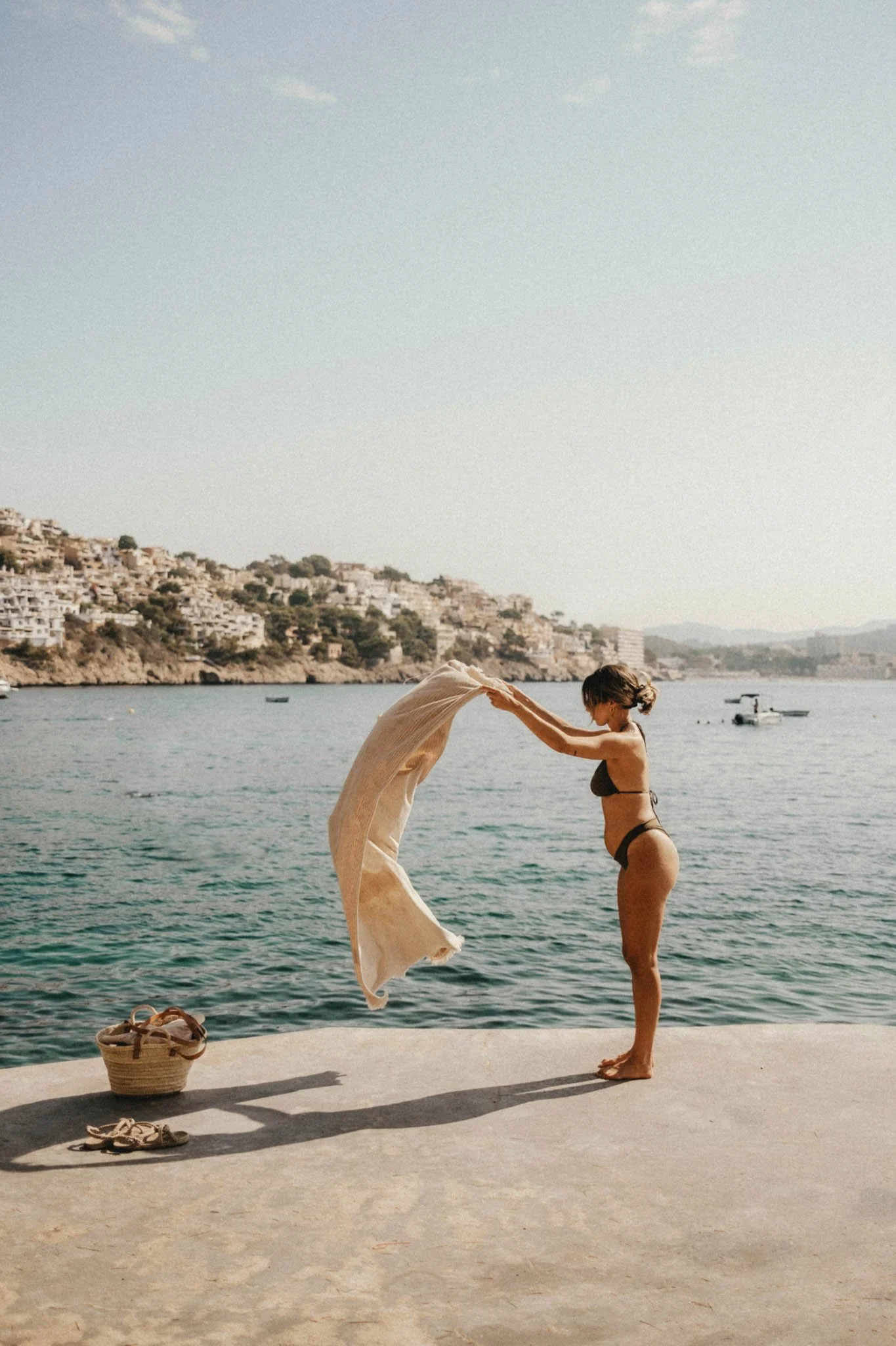 Une femme en maillot de bain noir épargne un vêtement blanc sur un quai près de la mer, avec des bateaux et des maisons en arrière-plan.