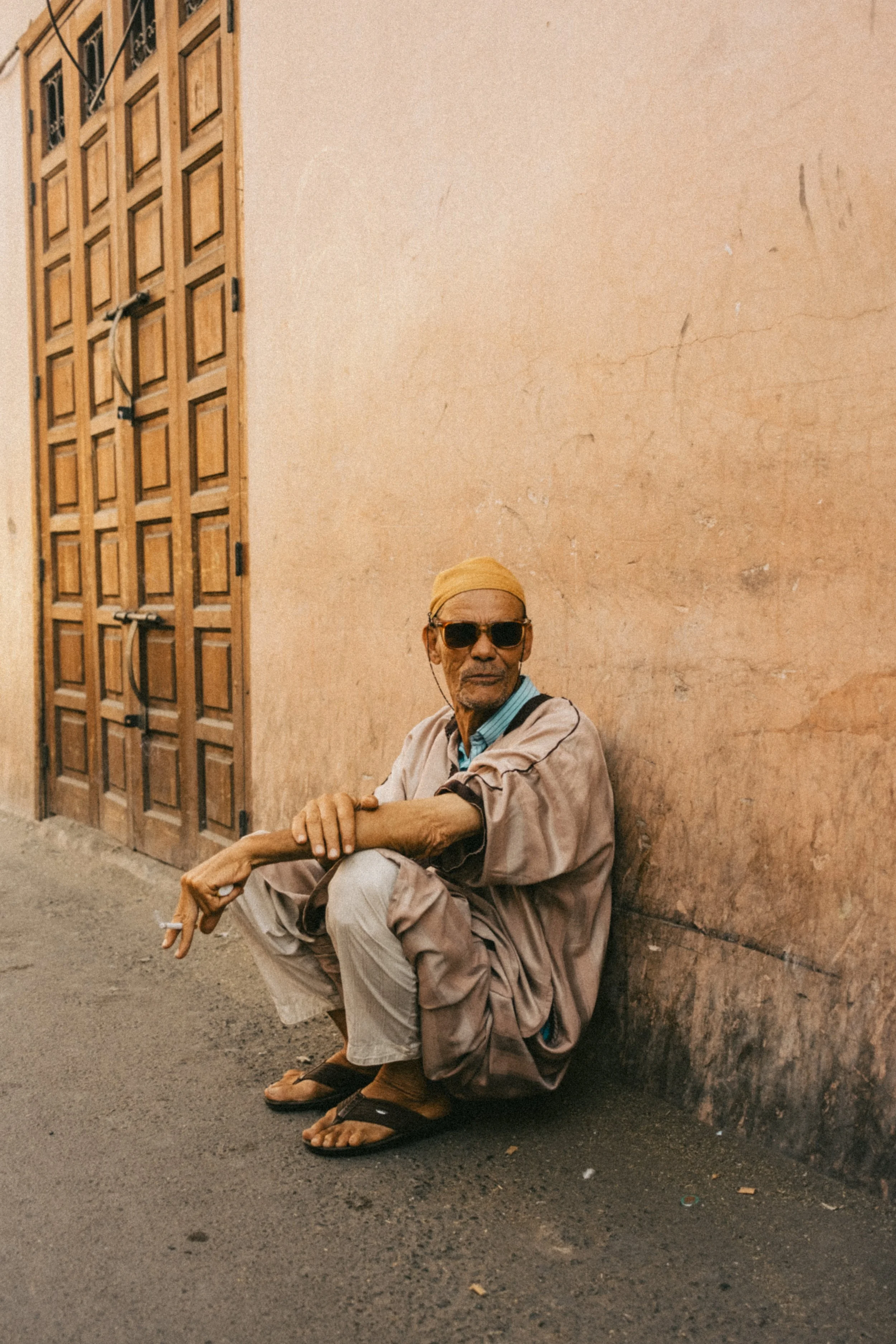 Un homme âgé portant des lunettes de soleil et un chapeau beige, assis contre un mur d'une couleur terre cuite, sur un trottoir en ville.