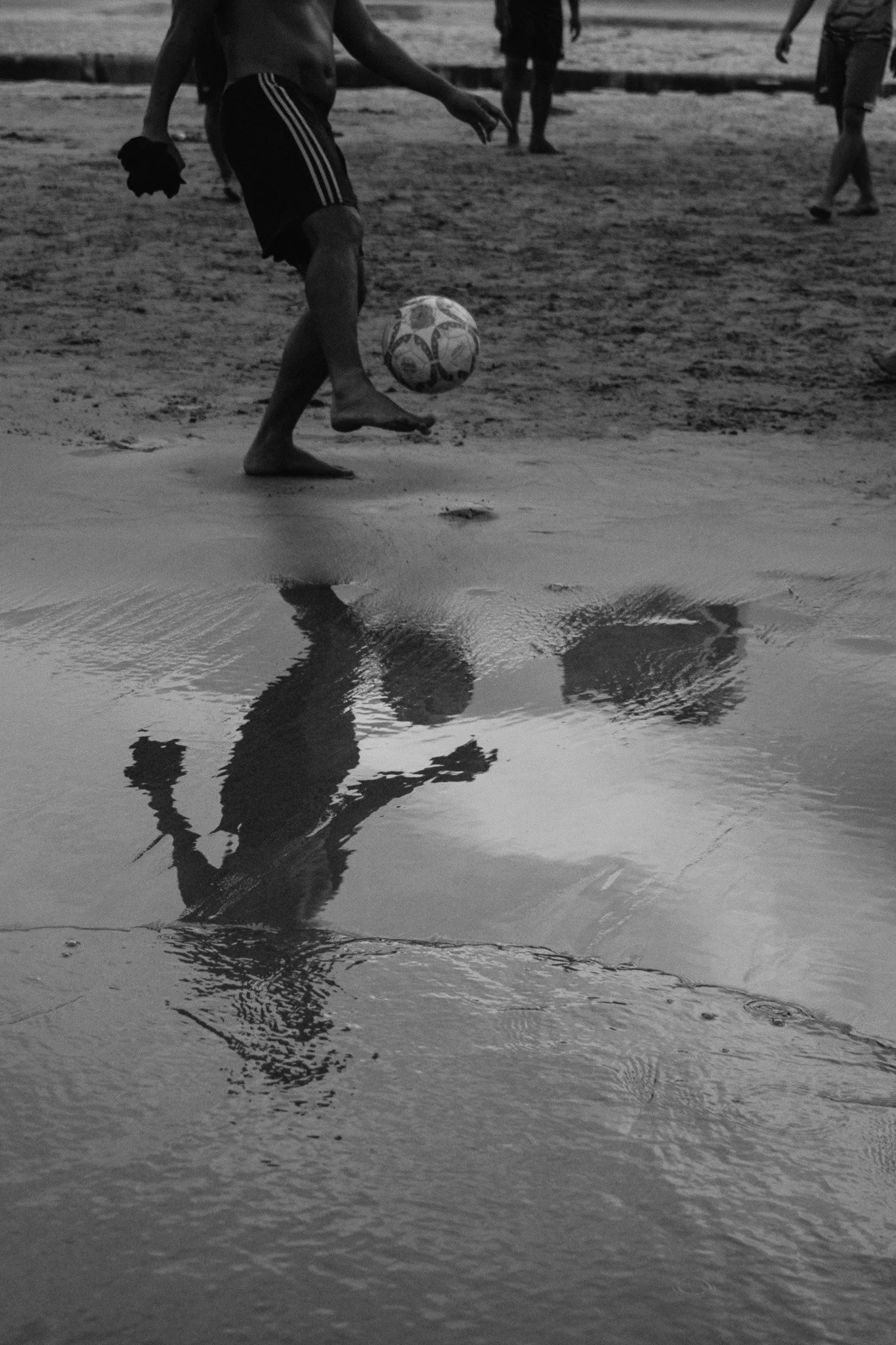 Un garçon joue au football sur la plage, sa réflexion apparaît dans une petite flaque d'eau à l'avant-plan, en noir et blanc.