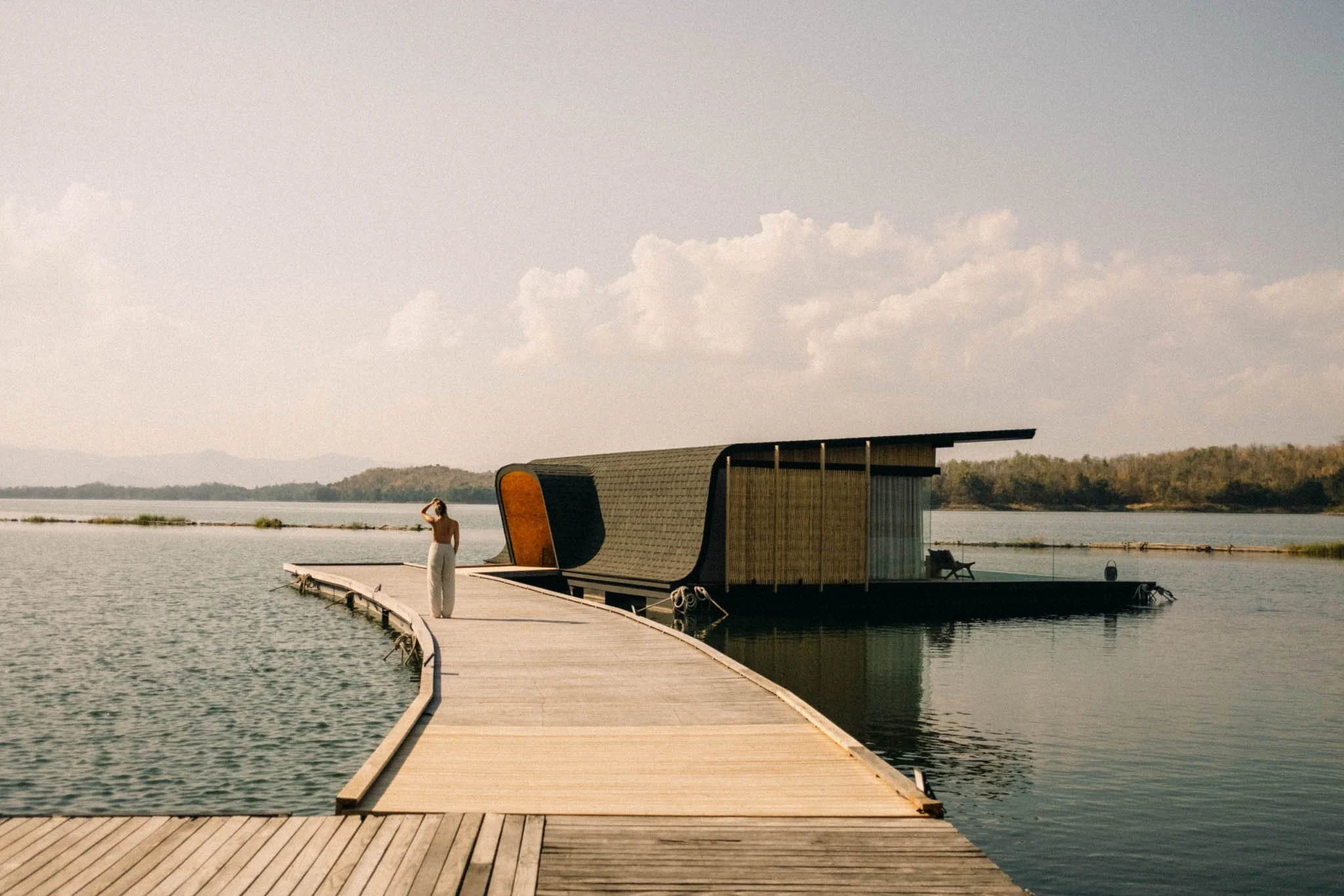 Une personne se tient sur un pont en bois menant à une maison flottante moderne sur un lac, avec un paysage naturel et un ciel nuageux en arrière-plan.