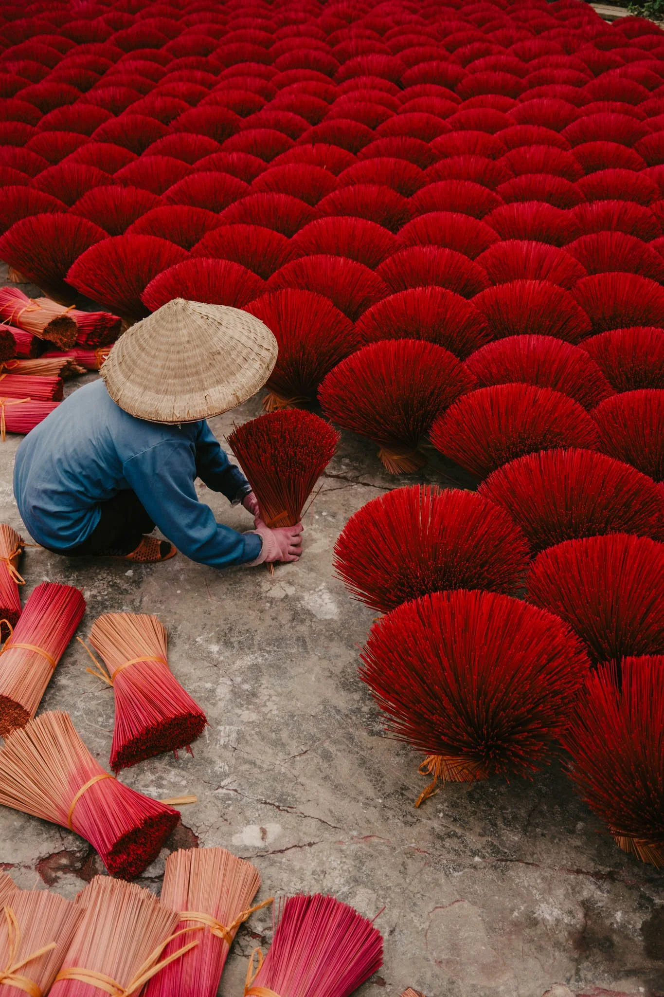 Une personne portant un chapeau conique en bambou prépare des éventails rouges en bambou sur un sol en béton.