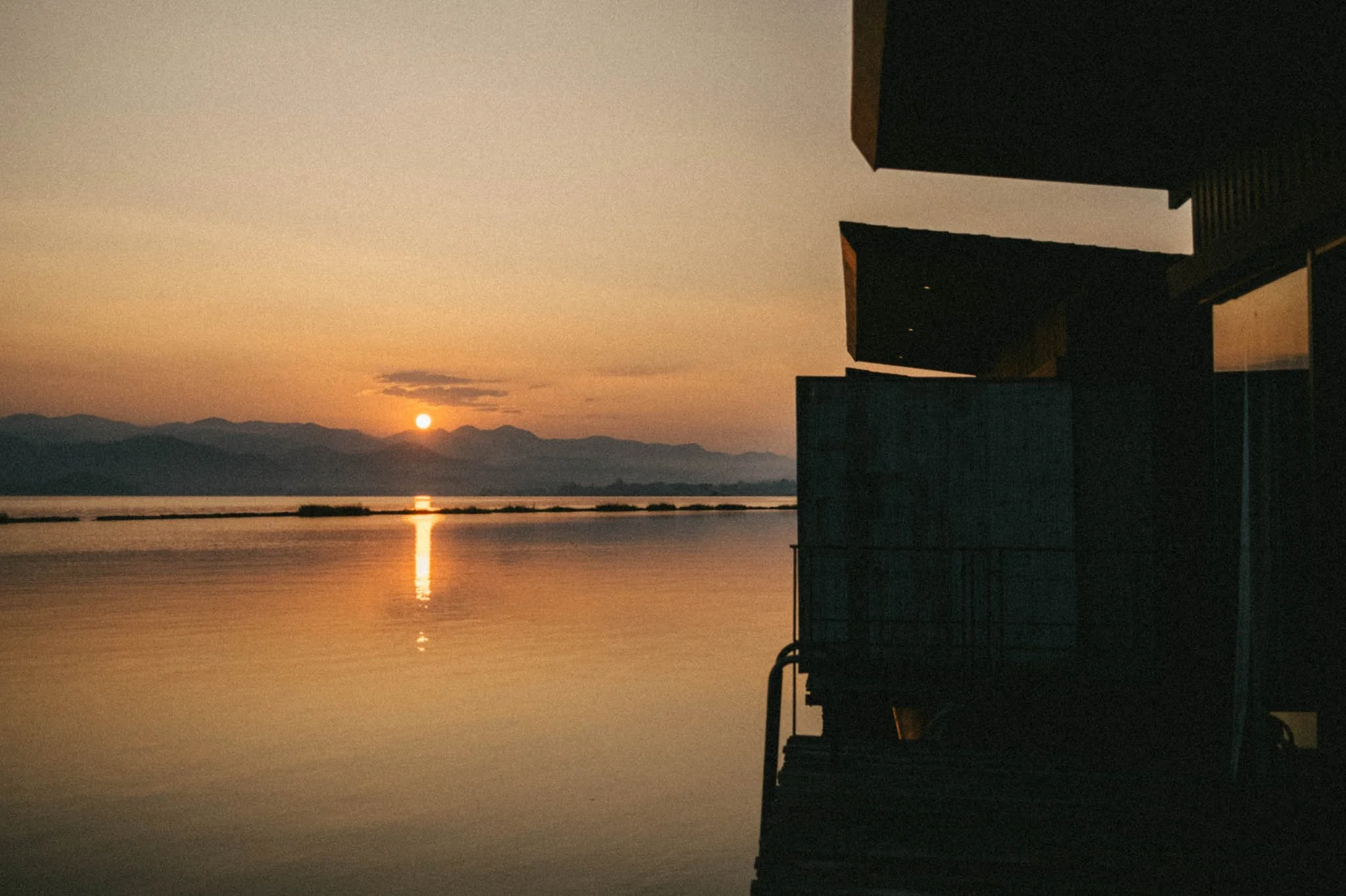 Coucher de soleil sur un lac avec montagnes en arrière-plan, vue partielle d'un bâtiment moderne en silhouette à droite.