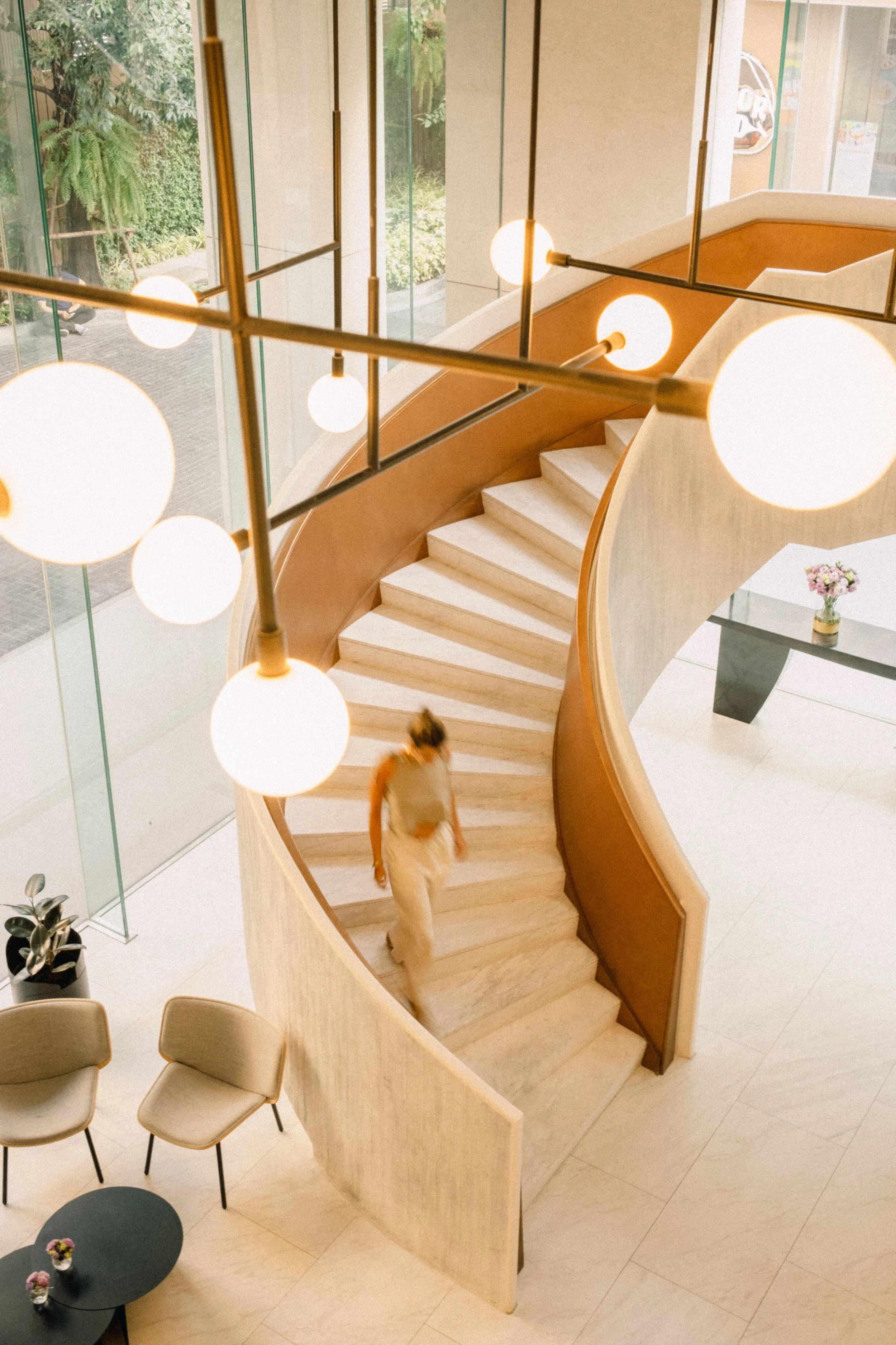 Intérieur d'un hall moderne avec escalier en colimaçon en bois clair portant une personne qui descend. Il y a une table avec des fleurs, un coin salon avec deux fauteuils et une petite table, et un lustre avec plusieurs globes lumineux suspendus au p