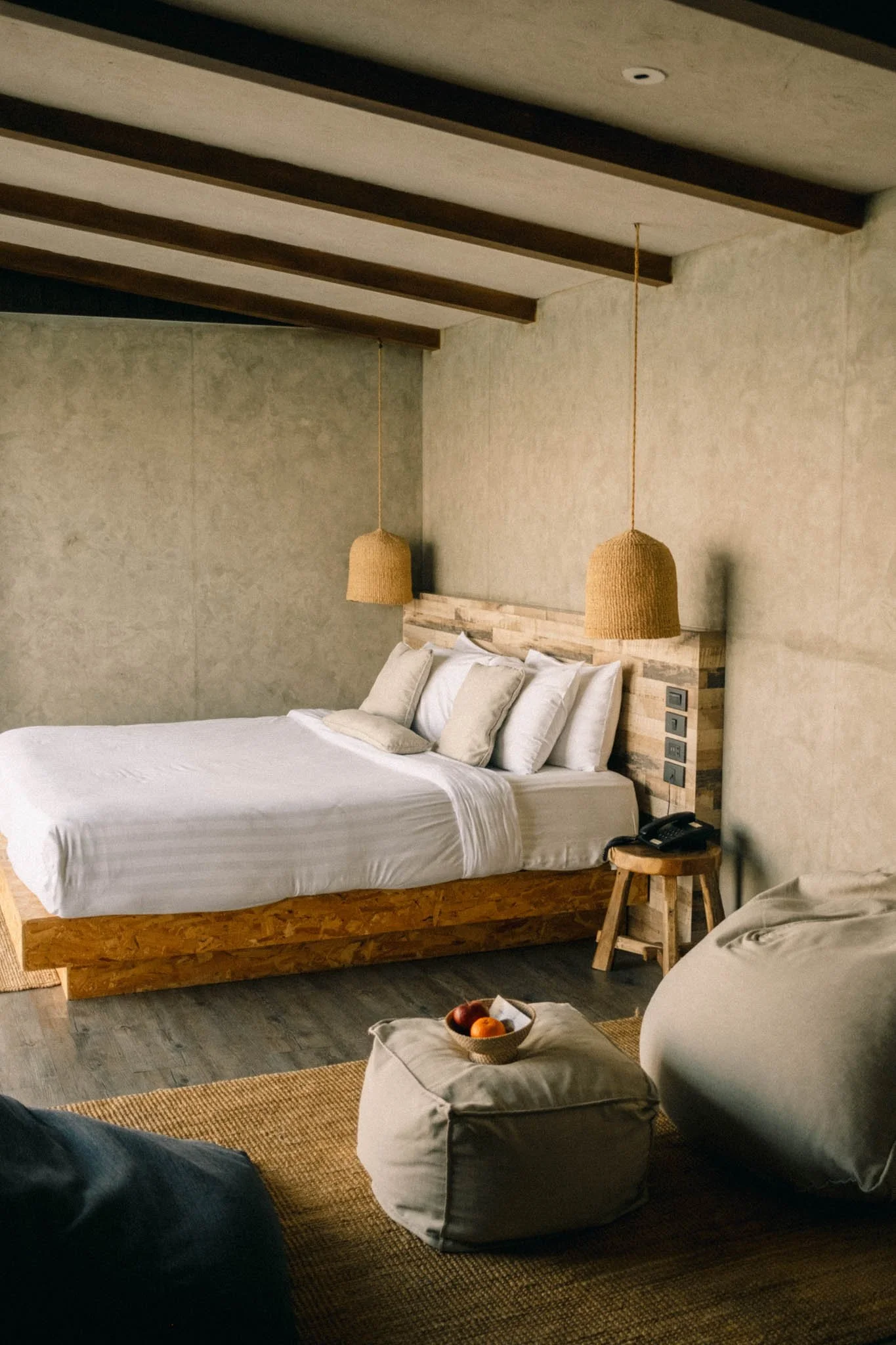 Chambre de style rustique avec un lit en bois, plusieurs oreillers blancs, deux suspensions en rotin, un téléphone sur une petite table en bois à côté du lit, et un pouf gris avec un bol de fruits, sur un tapis en jonc de mer.