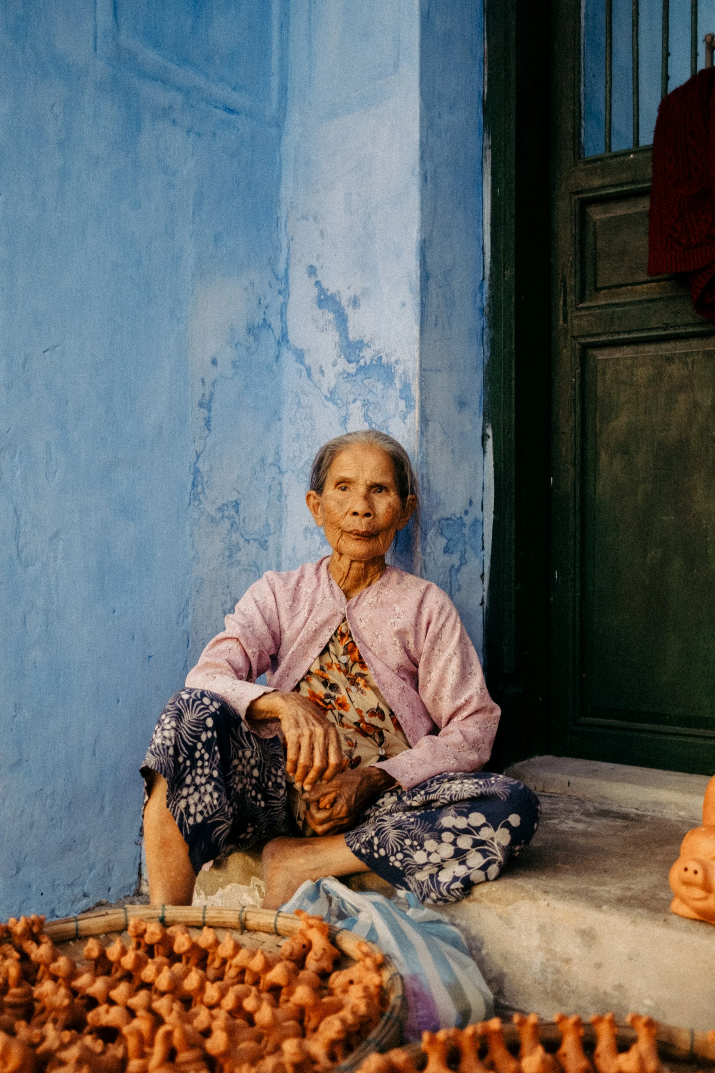 Femme âgée assise sur le sol devant un mur bleu, avec des objets en argile autour d'elle.