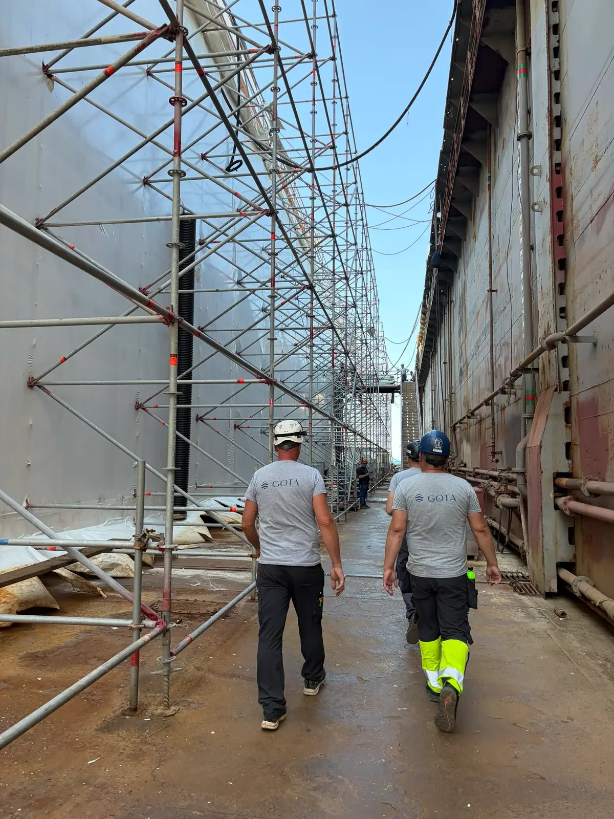 Construction workers walking through a site with scaffolding on one side and a weathered building wall on the other, under blue sky in the background.