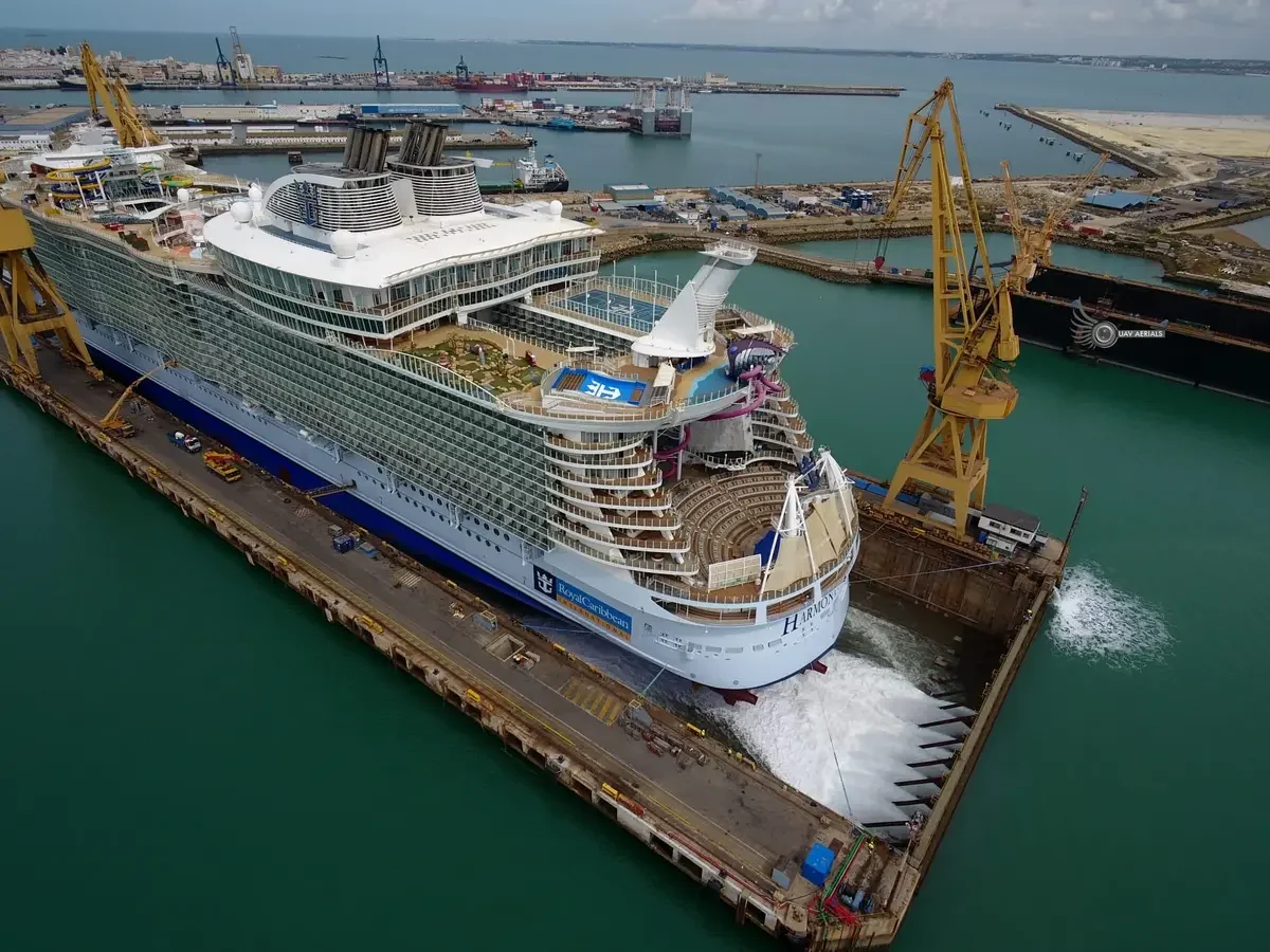A large cruise ship named Harmony docked at a harbor under construction, with cranes and construction equipment around it, and water surrounding the port.