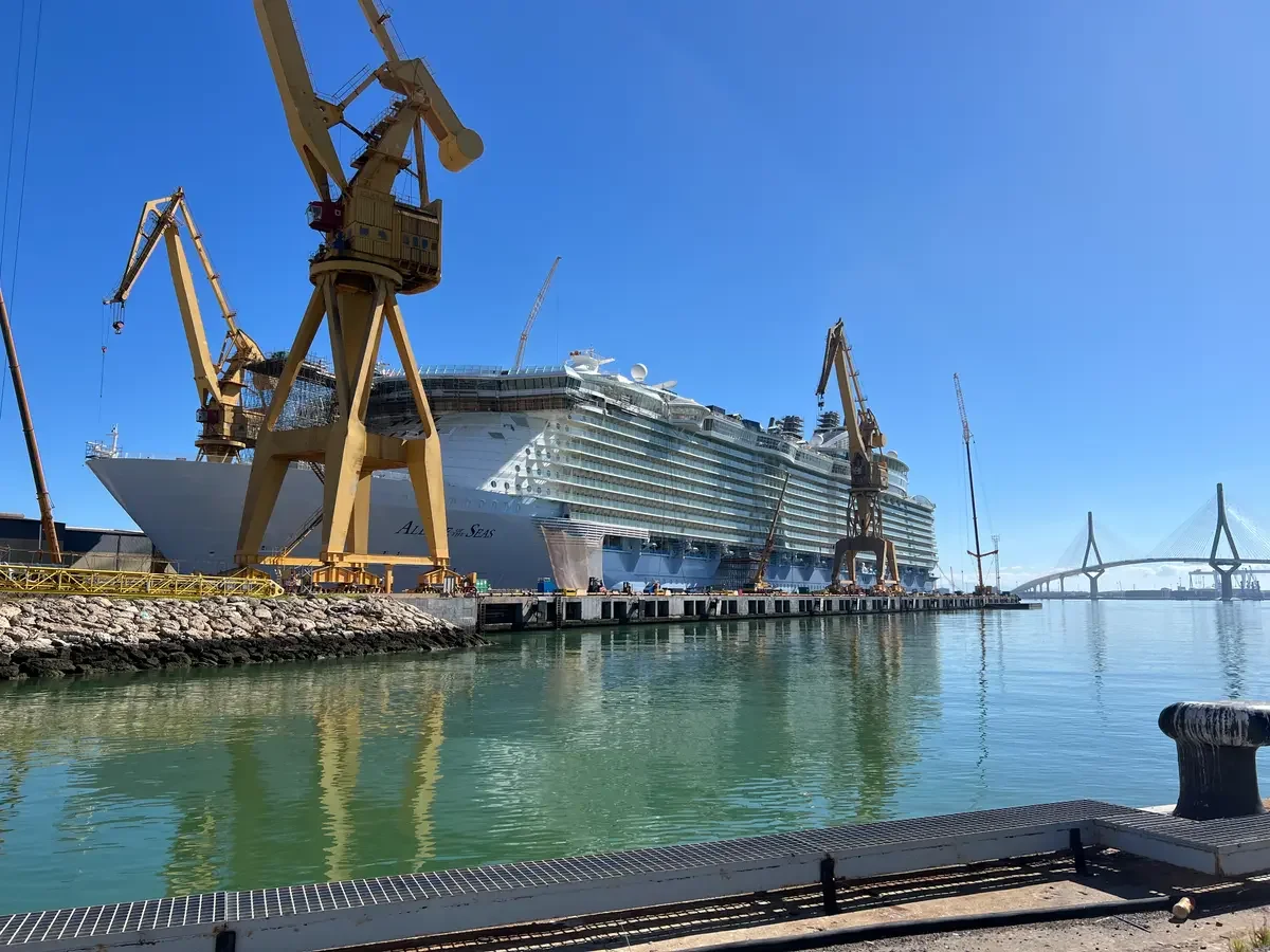 Large cruise ship docked at a port, surrounded by large yellow construction cranes, with a bridge in the background, and calm green water in the foreground.