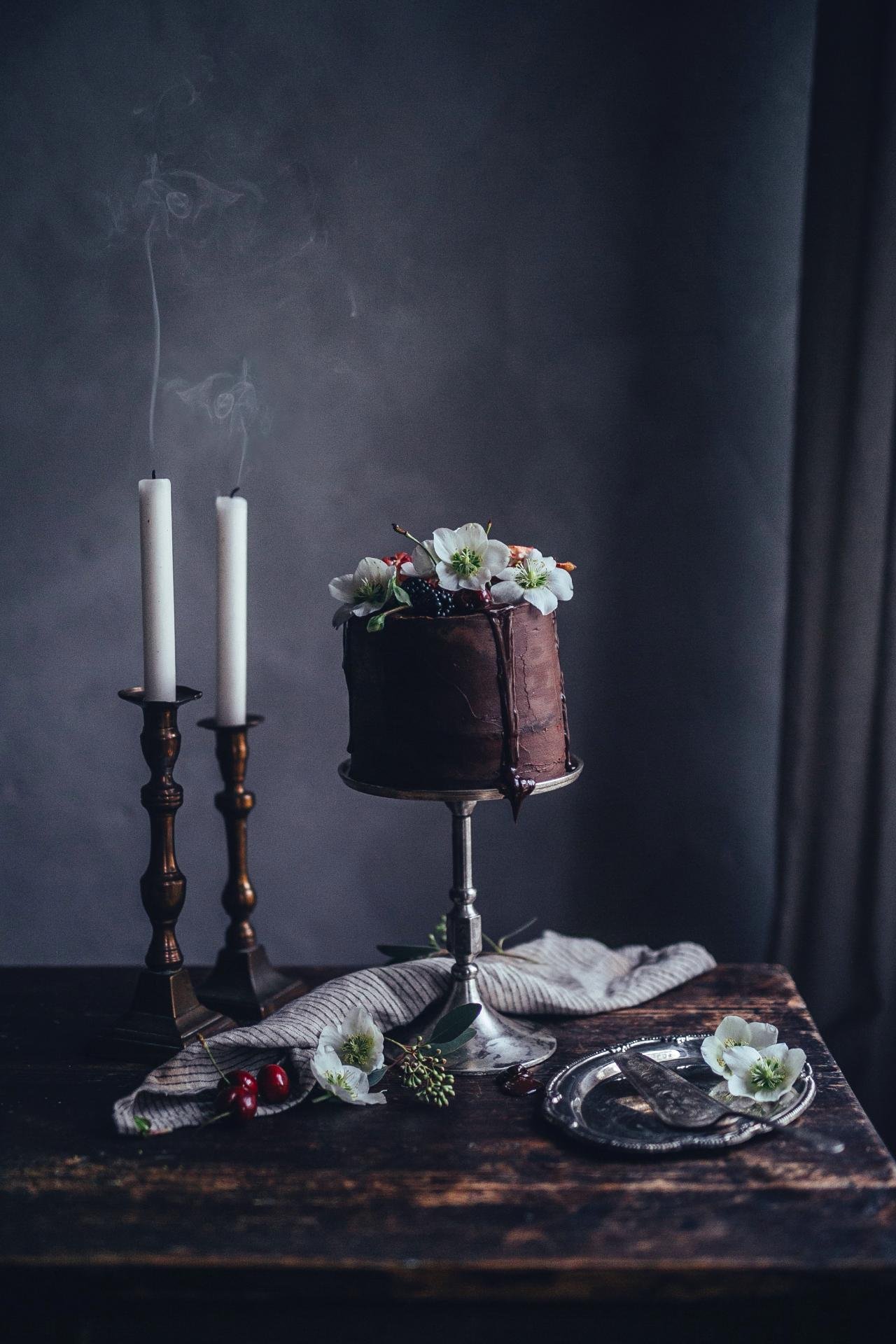 a beautiful chocolate birthday cake decorated with fresh fruits and flowers displayed on a wooden table with candles and silver tray