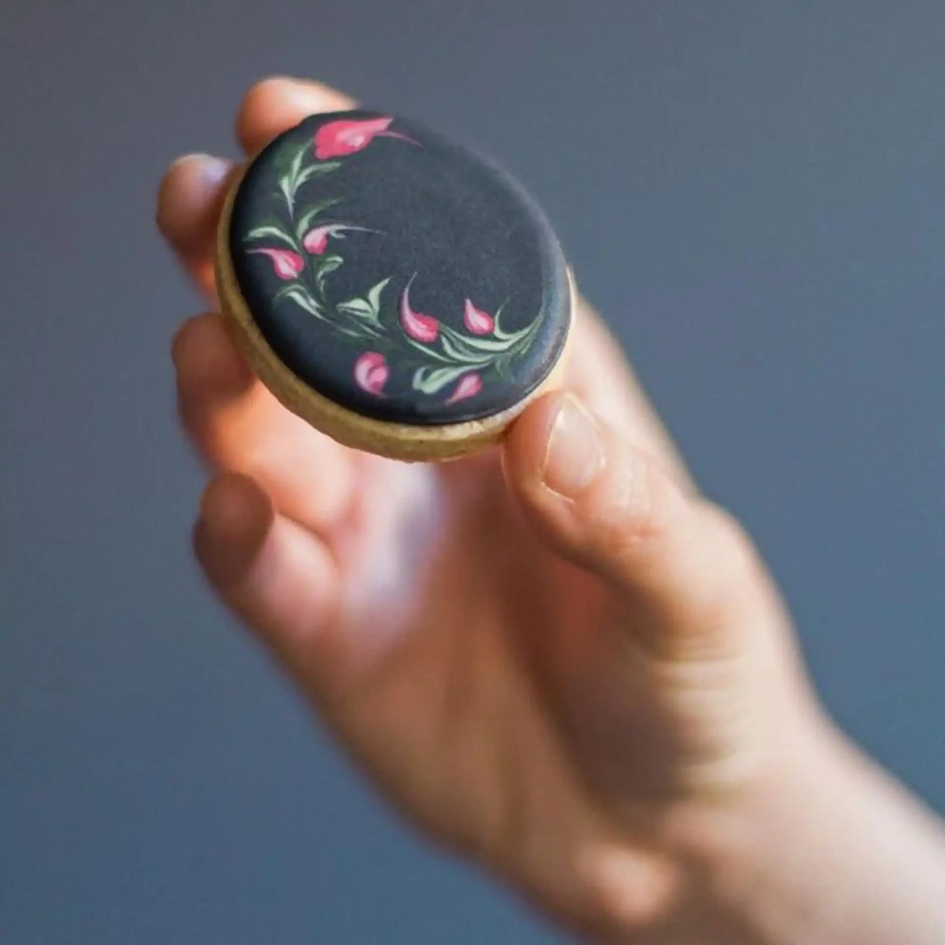 A hand holding a round, black object decorated with a floral design featuring pink flowers and green leaves.