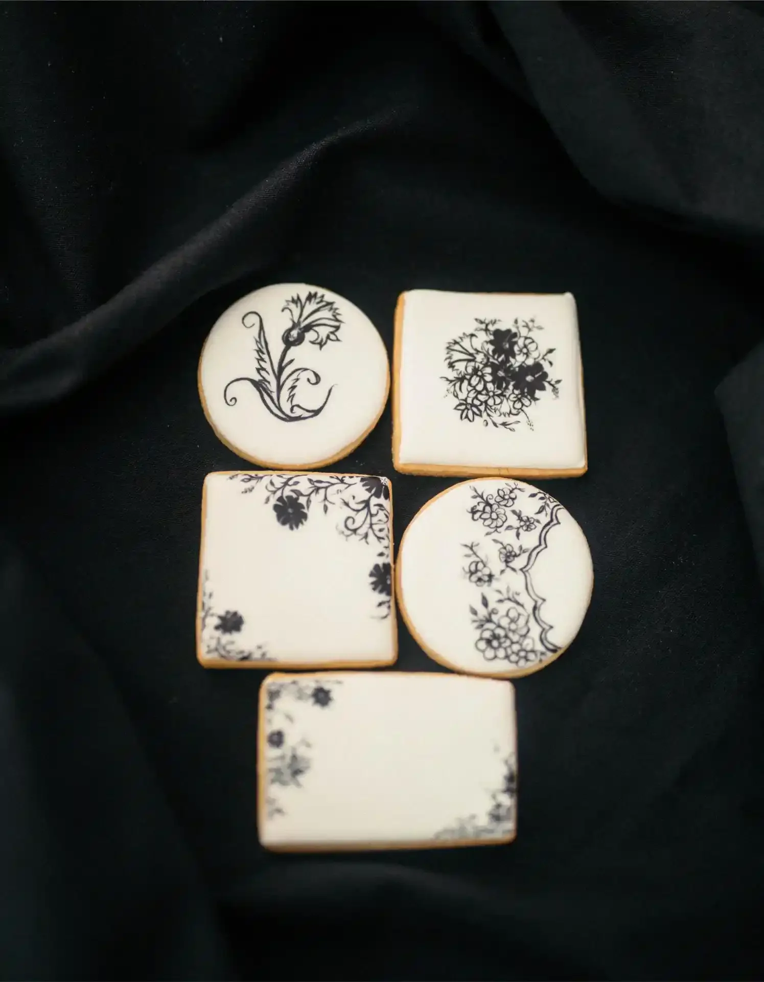 Set of decorative cookies with black floral and botanical patterns on white icing, placed on black fabric.