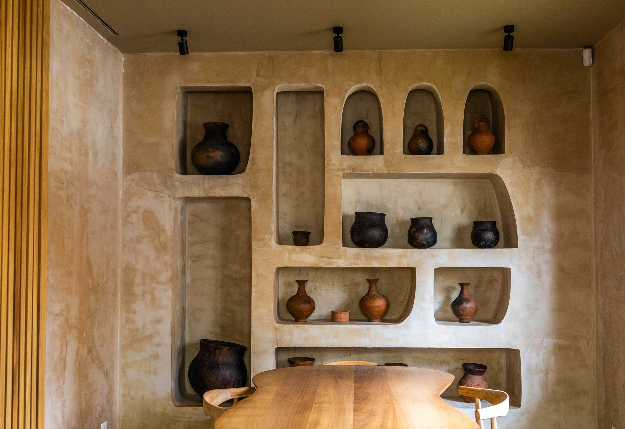 Estante embutida na parede com várias argolas de cerâmica e barro em tons escuros e marrons, decorativa em um ambiente interno, com uma mesa de madeira ao fundo.