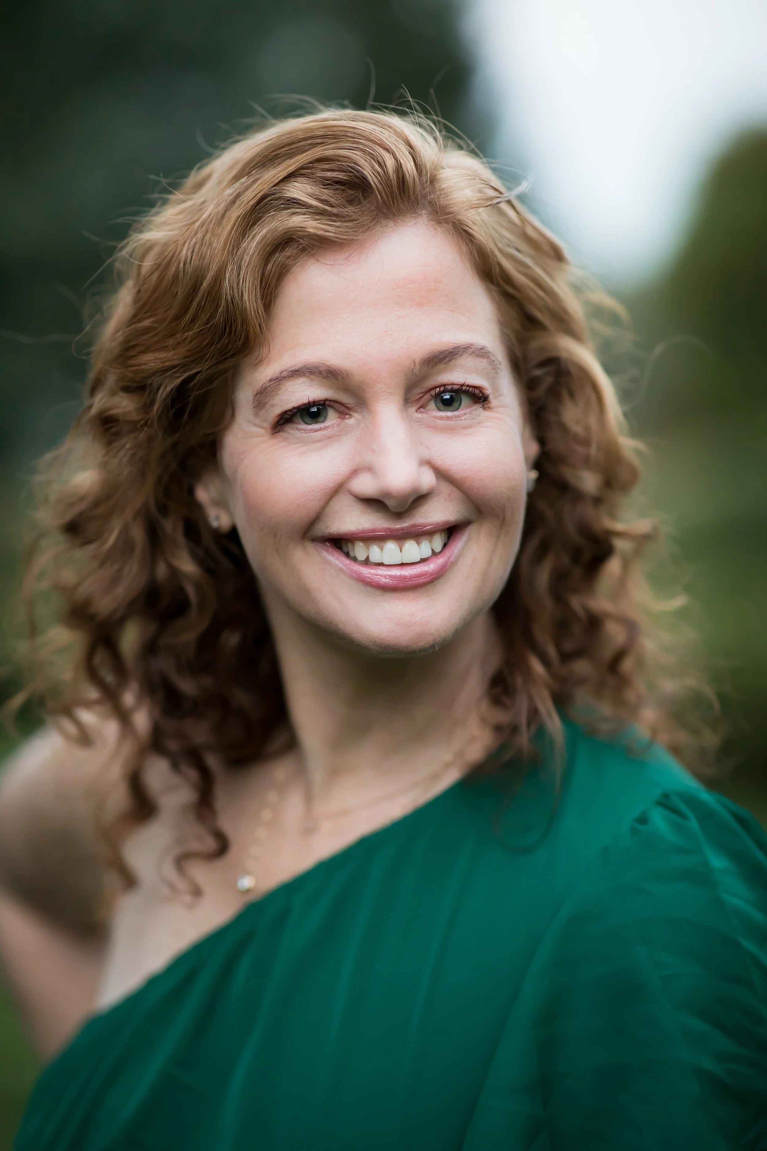 Close-up of a woman with curly red hair, smiling outdoors, wearing a green dress and a delicate necklace.