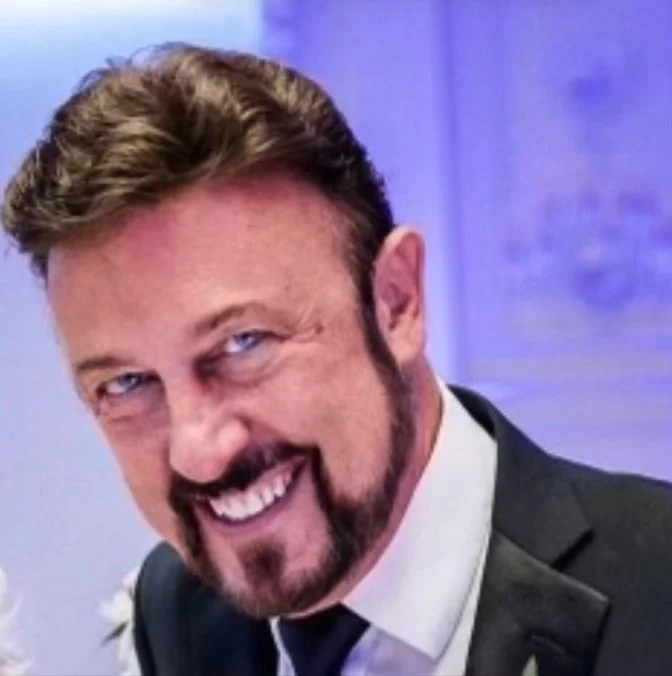 Close-up of a man with a mischievous expression, smirking, with short brown hair, a beard, and wearing a dark suit and tie.
