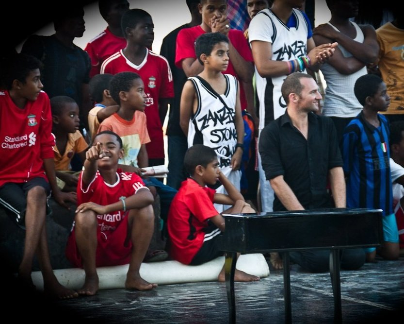 Group of children and a man with light skin, seated and standing together, some wearing sports jerseys, in an indoor setting.