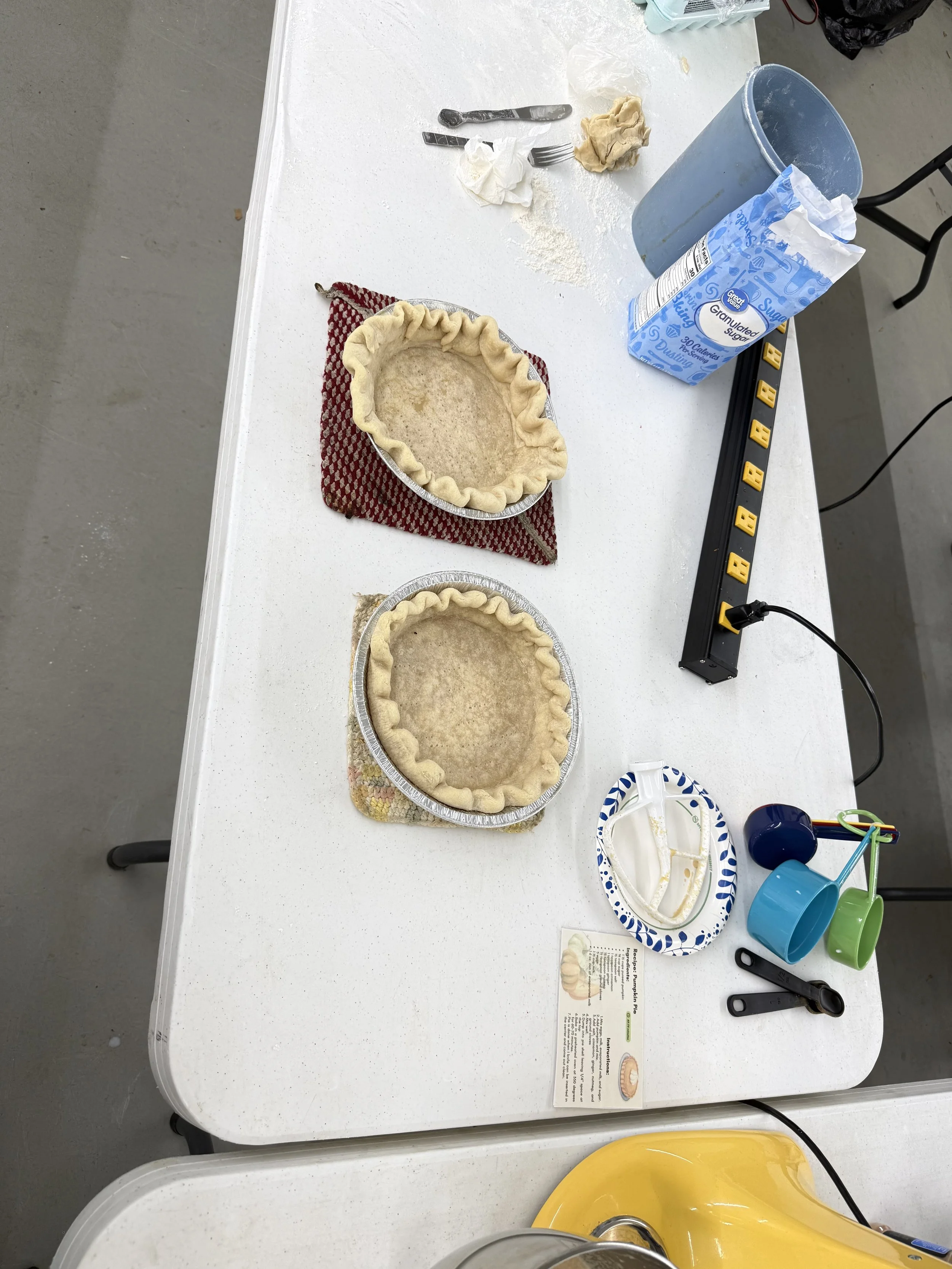 Unfinished pie crust in aluminum baking pans on a white table, with baking tools, sugar, and a recipe card nearby.