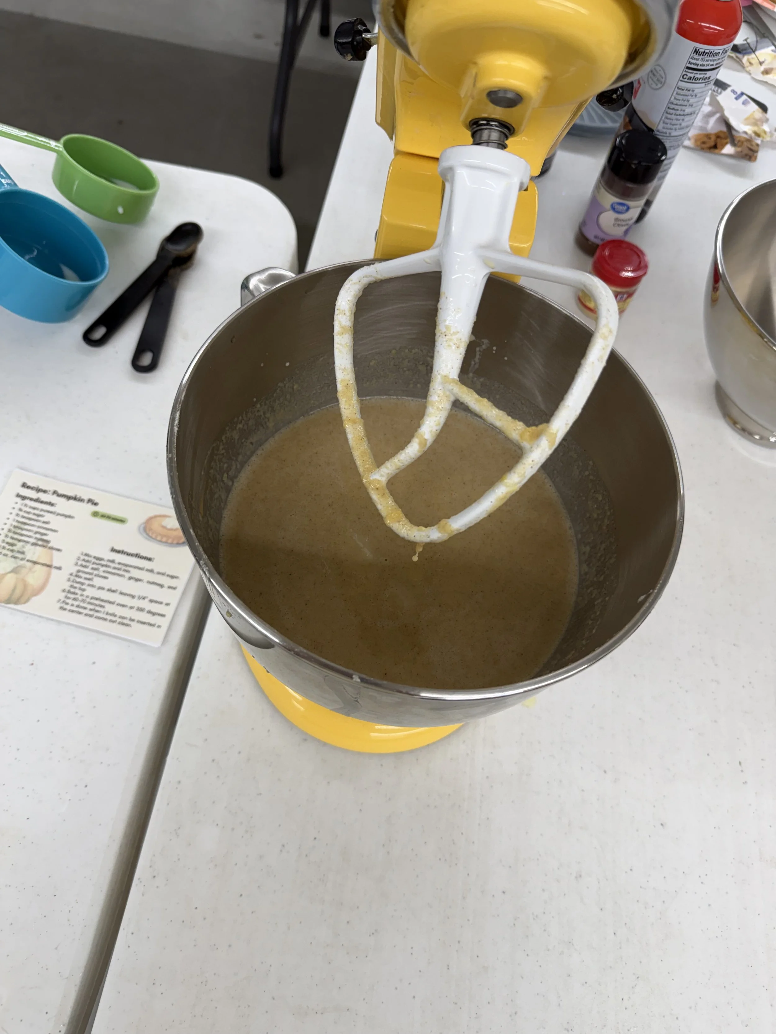 A stand mixer is mixing what looks like pumpkin pie filling in a metal bowl on a kitchen counter. There are various baking ingredients and tools visible on the countertop.