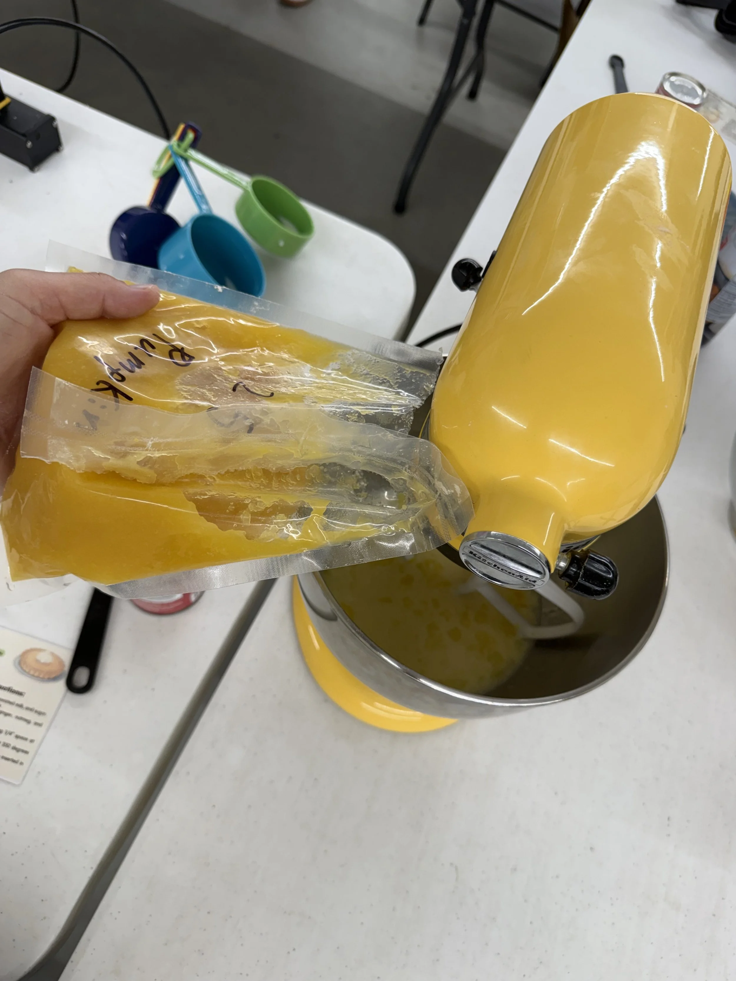 A yellow kitchen appliance with a foam layer on top, a person's hand holding a vacuum-sealed packet of yellow substance, on a white countertop with colorful cups in the background.