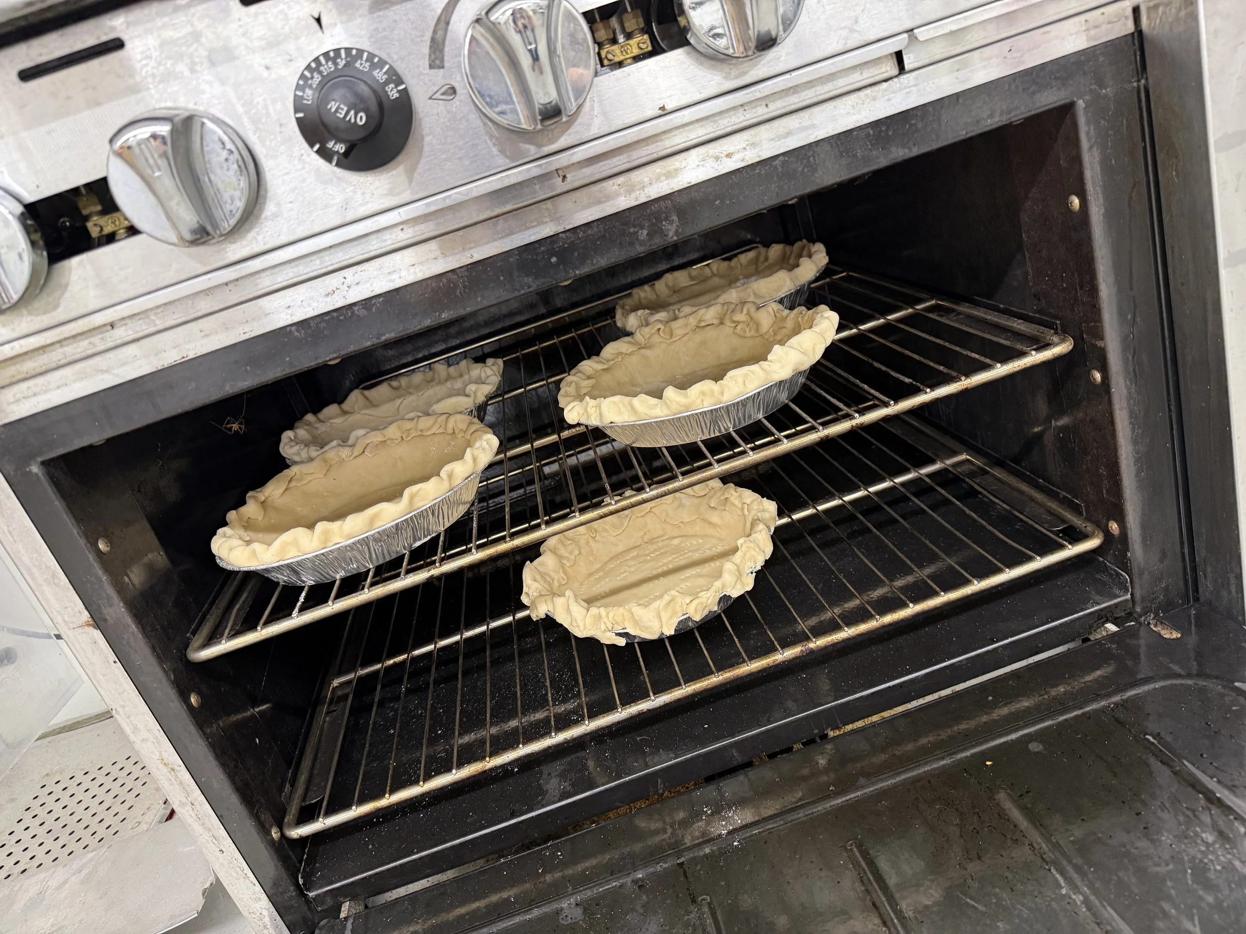 Oven with six pie crusts in aluminum pans baking on oven racks.