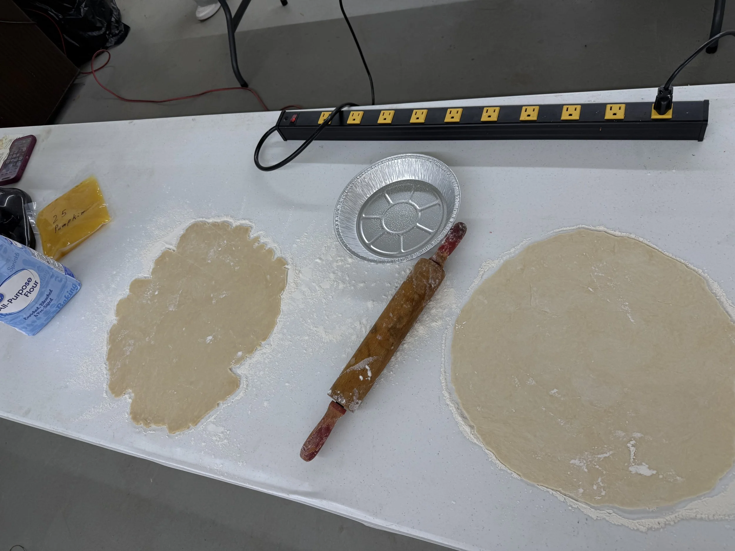 A white countertop with rolled-out dough, a rolling pin, a foil tray, a package of flour, and a power strip with multiple outlets.