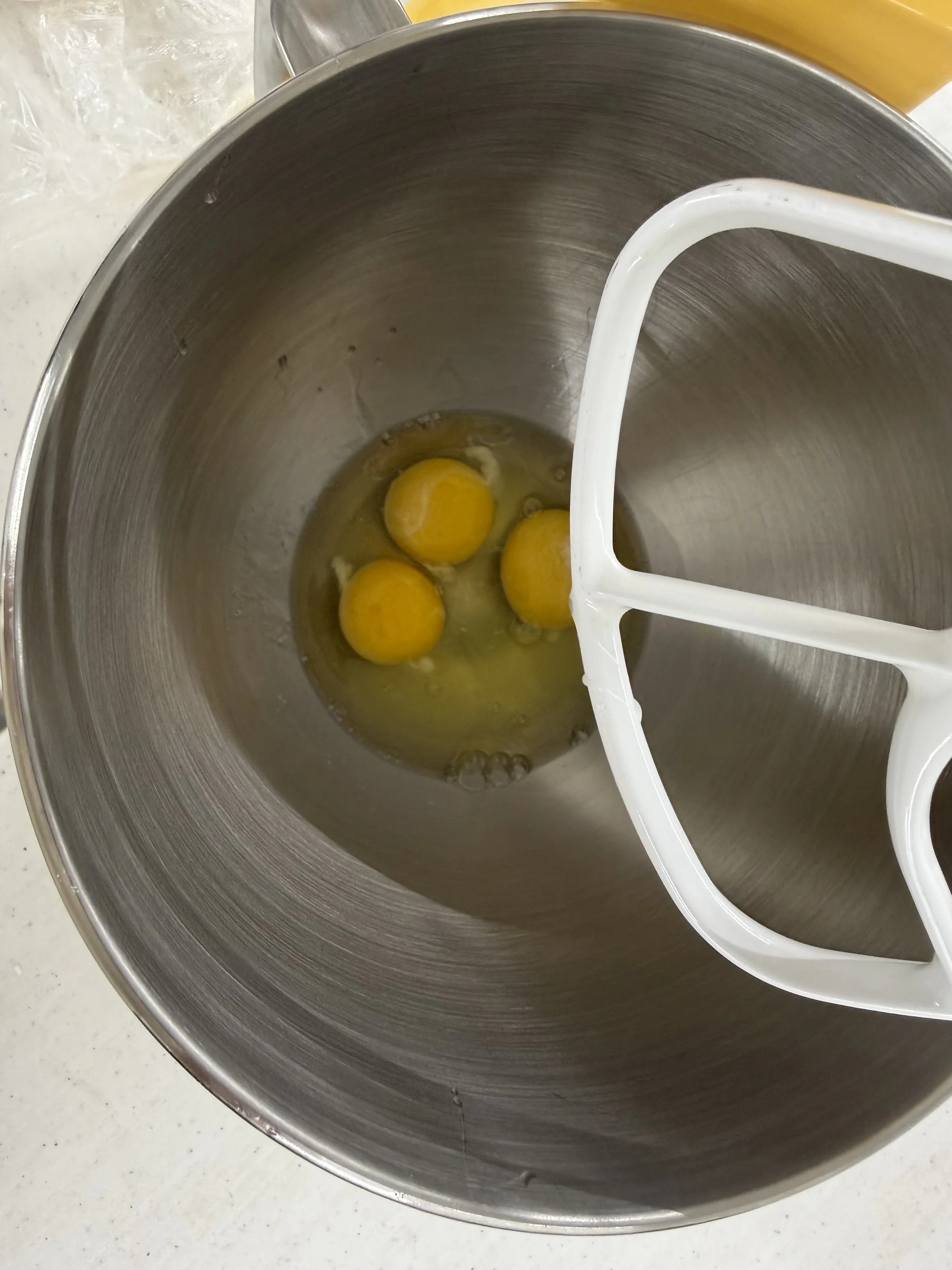 A stand mixer with three eggs inside. The mixer has a white beaters attachment and is placed on a white countertop.