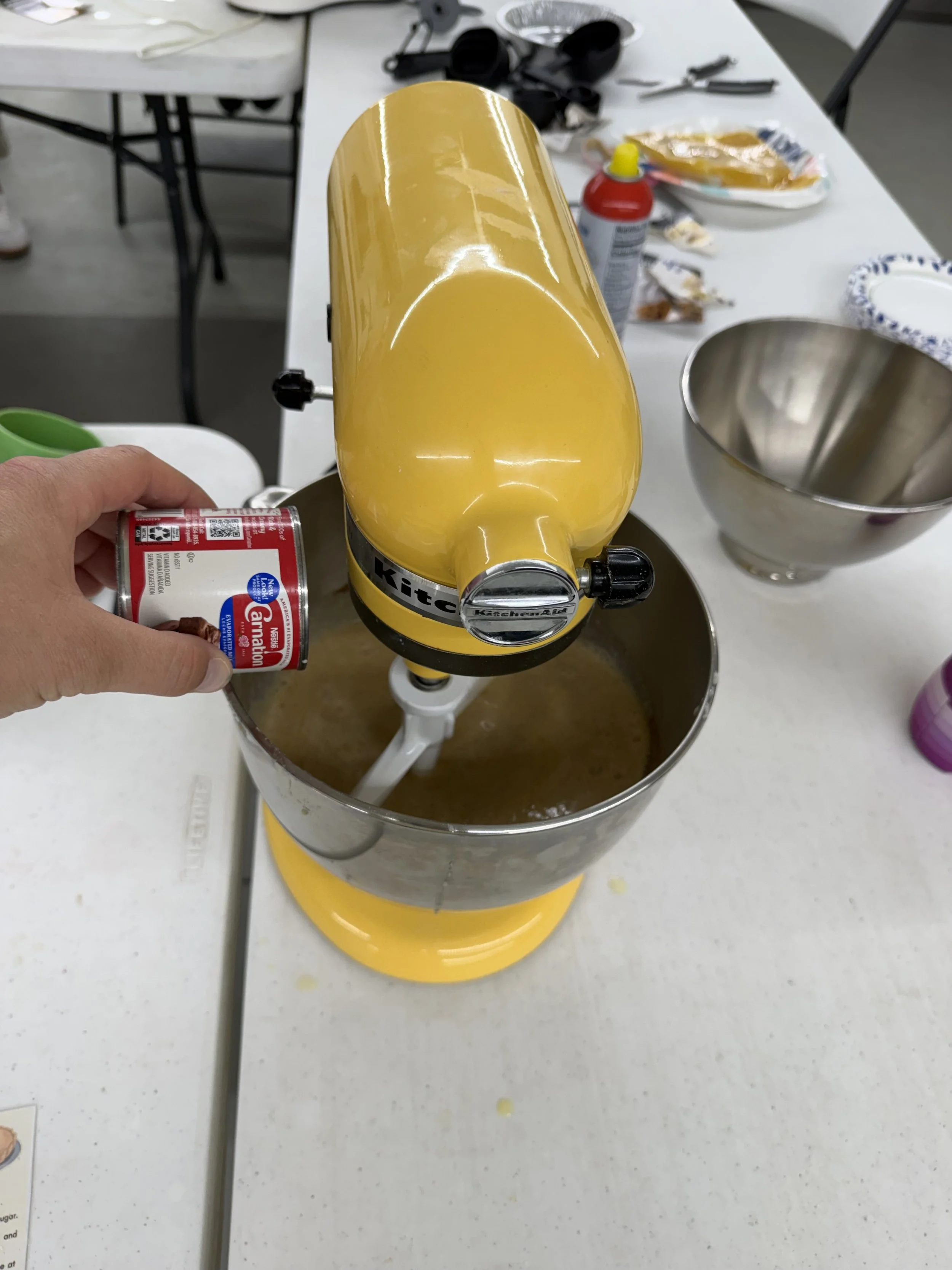 Close-up of a yellow KitchenAid stand mixer with a bowl underneath, in a kitchen or workspace, with various kitchen tools and ingredients on the table around it.