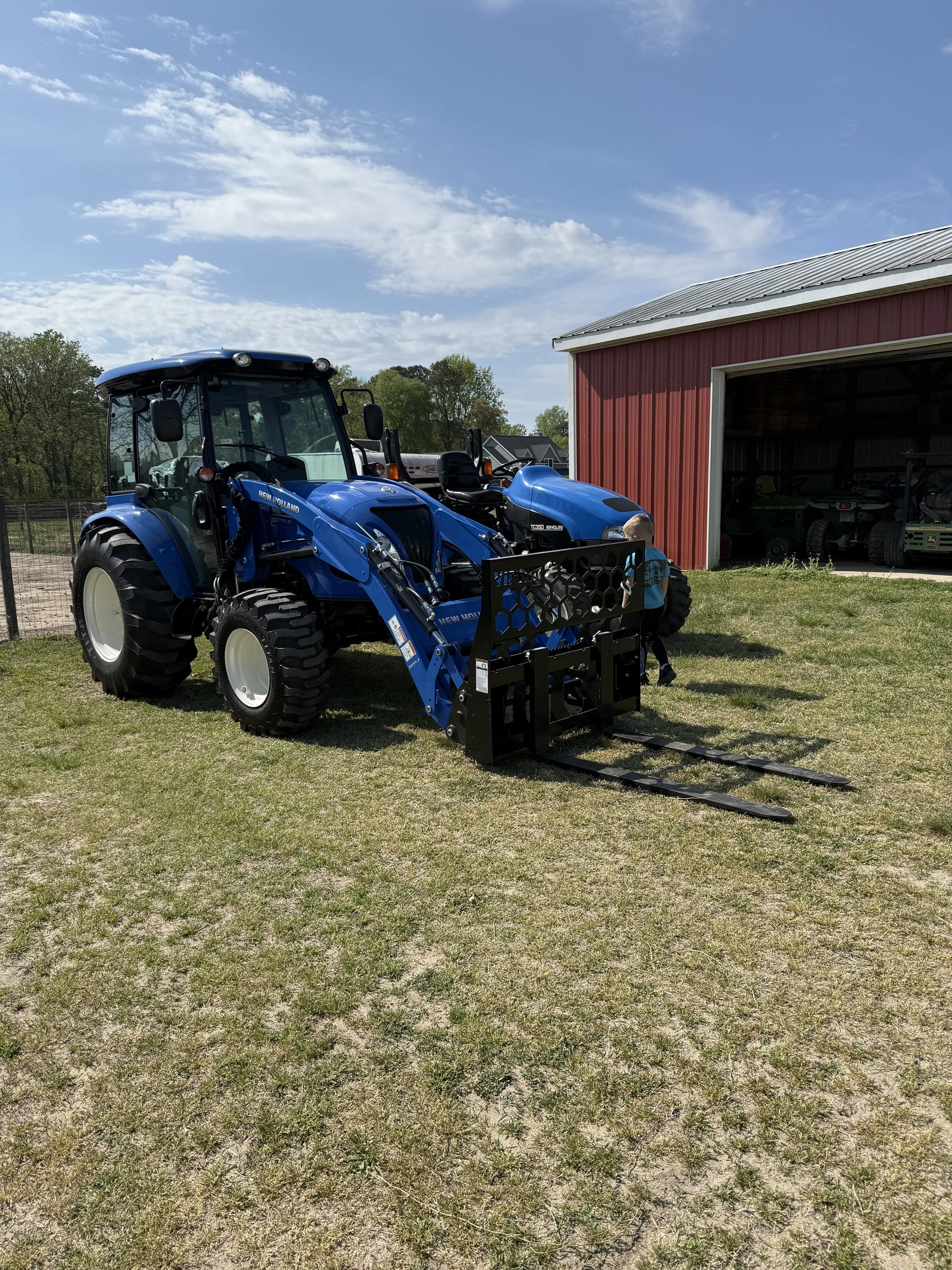 A blue New Holland tractor parked outside a red barn with an open door, on a grassy area under a blue sky with scattered clouds.