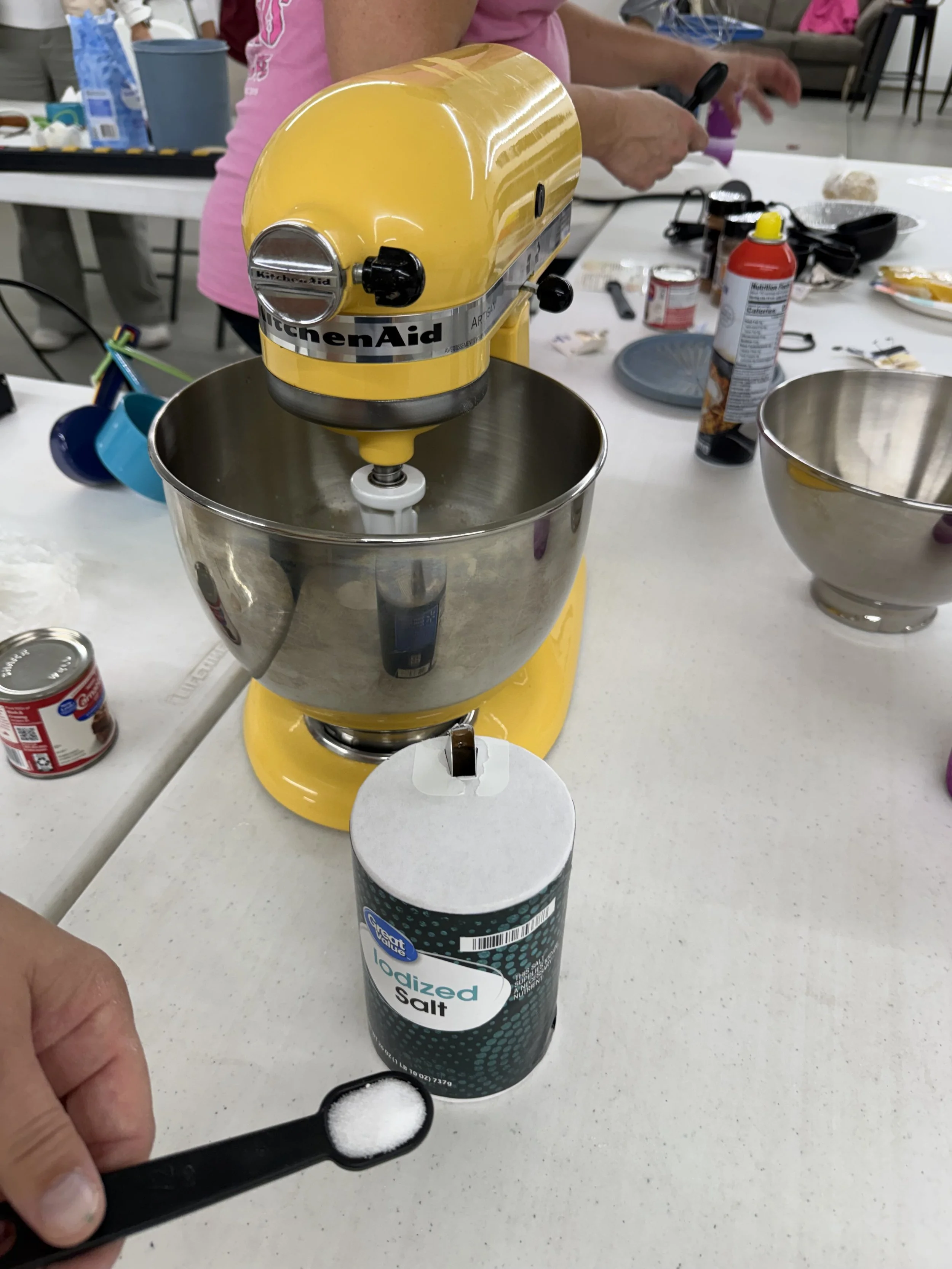 A yellow KitchenAid stand mixer with a mixing bowl attached, on a table with various baking ingredients and tools. A hand is holding a black measuring spoon with salt over the salt container.