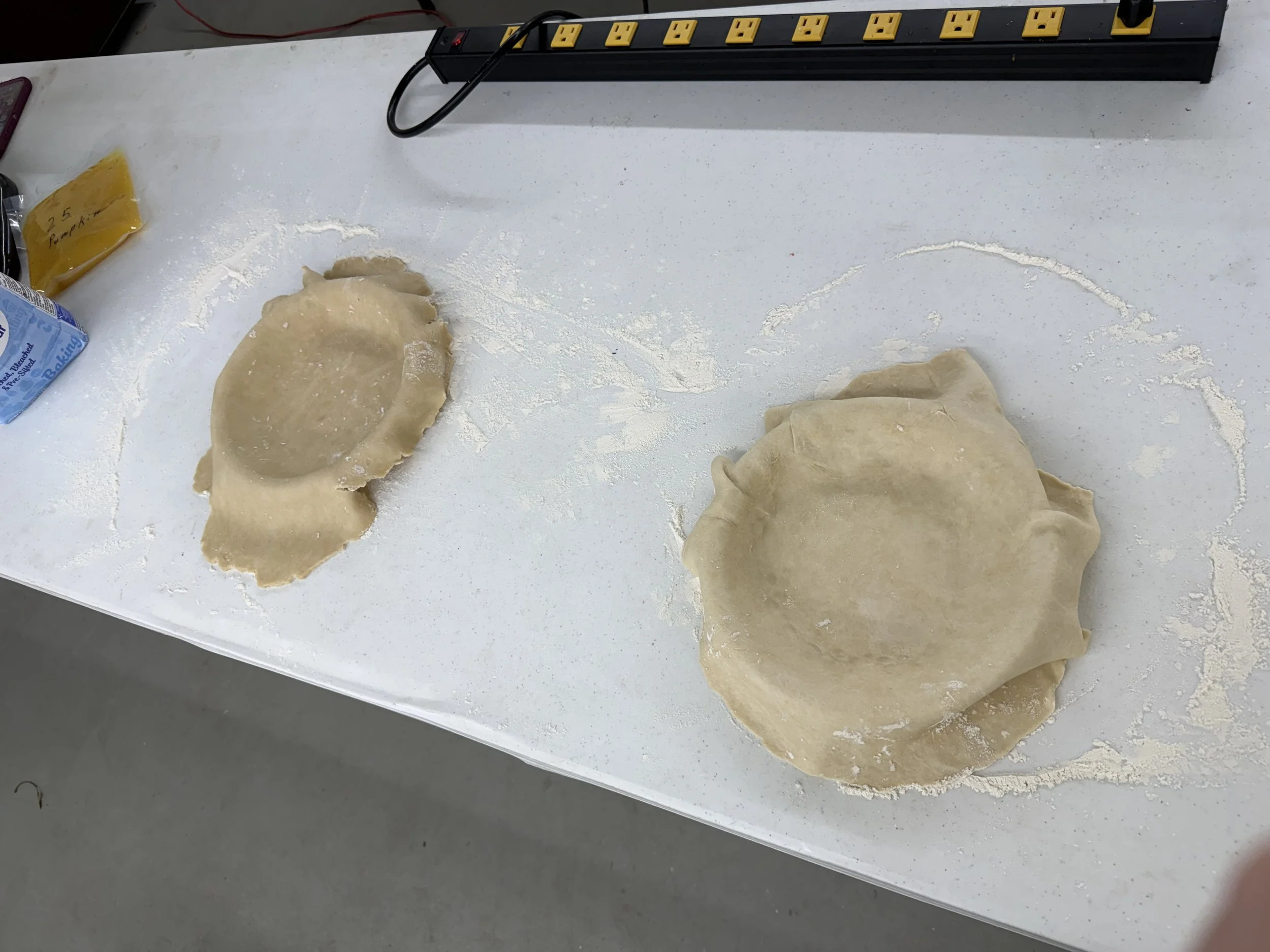 Unrolled disc of pie dough on a shellacked white countertop with a small pile of extra dough to the right. There is flour scattered on the surface and a plastic bag of pre-sliced butter with blue packaging on the left, and a yellow note on the corner