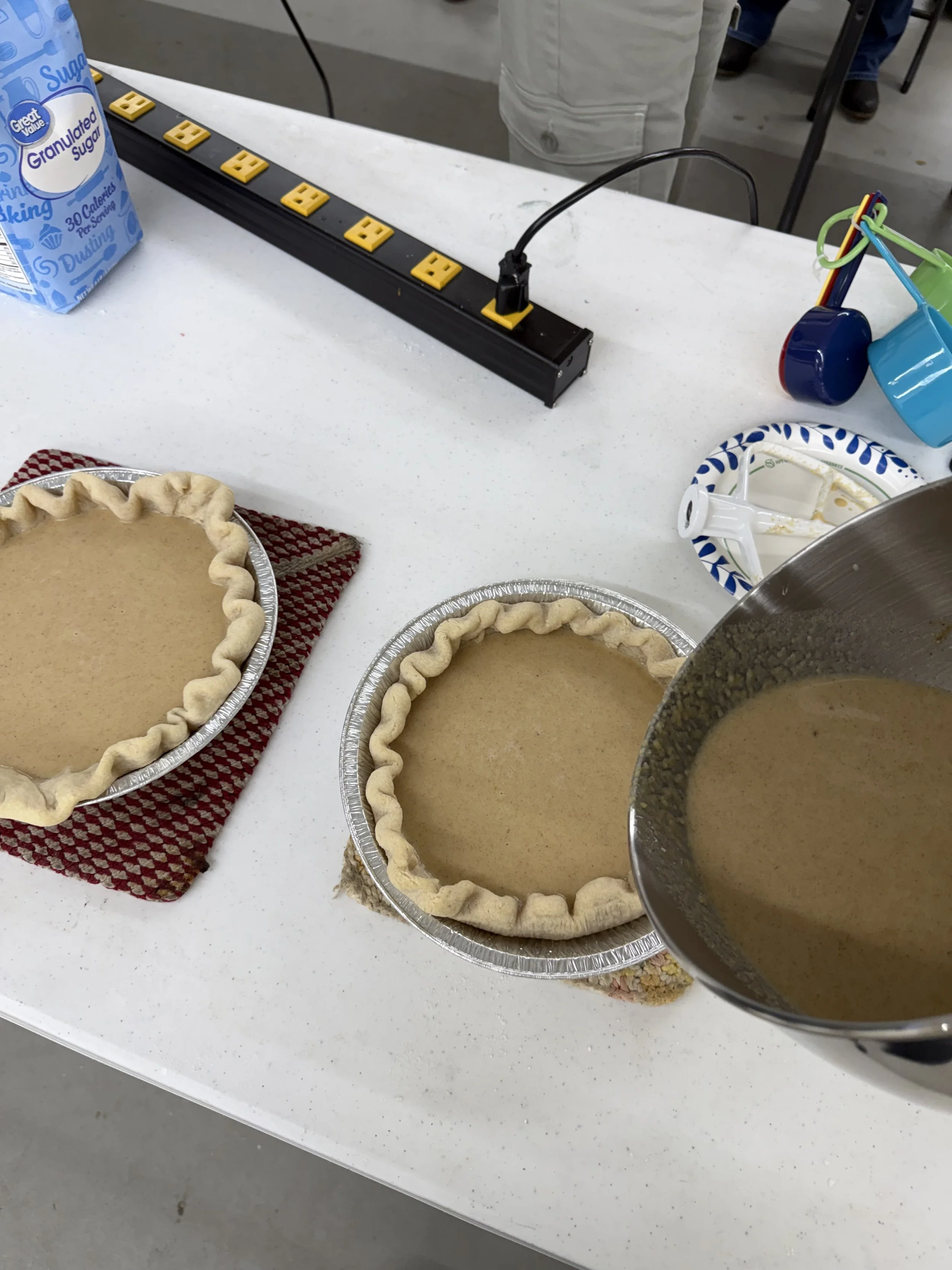 Three aluminum pies with unbaked crusts on a white table, with one metal bowl containing a liquid filling being poured into one pie. In the background, there is a carton of granulated sugar, colored mugs, and a cloth.