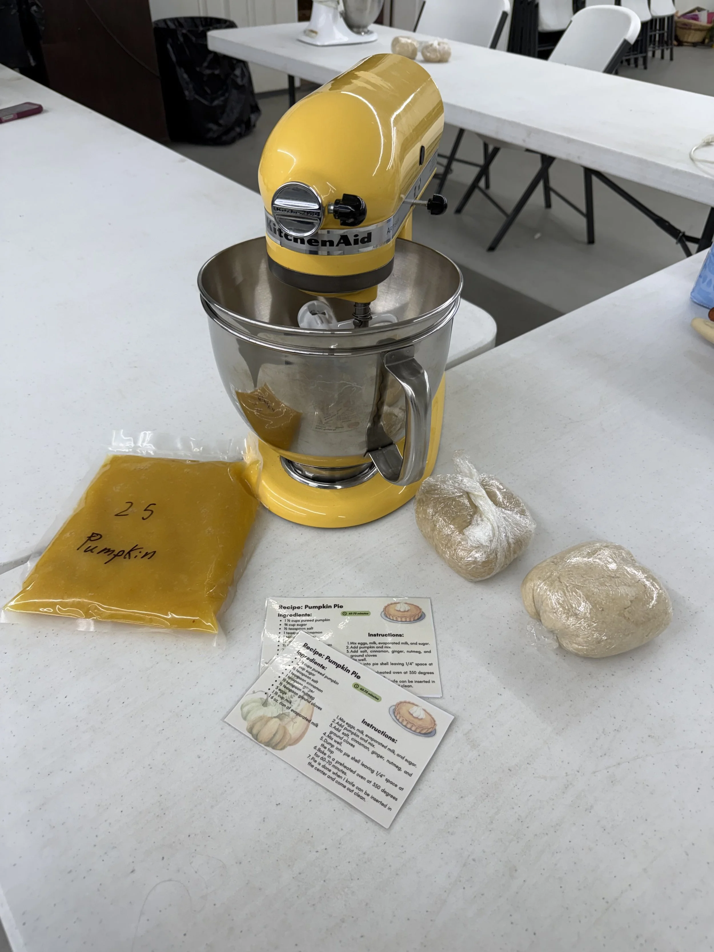 A yellow KitchenAid stand mixer attached to a mixing bowl, with a bag of pumpkin puree labeled "25 Pumpkin" and some dough balls and a recipe card on a white countertop.