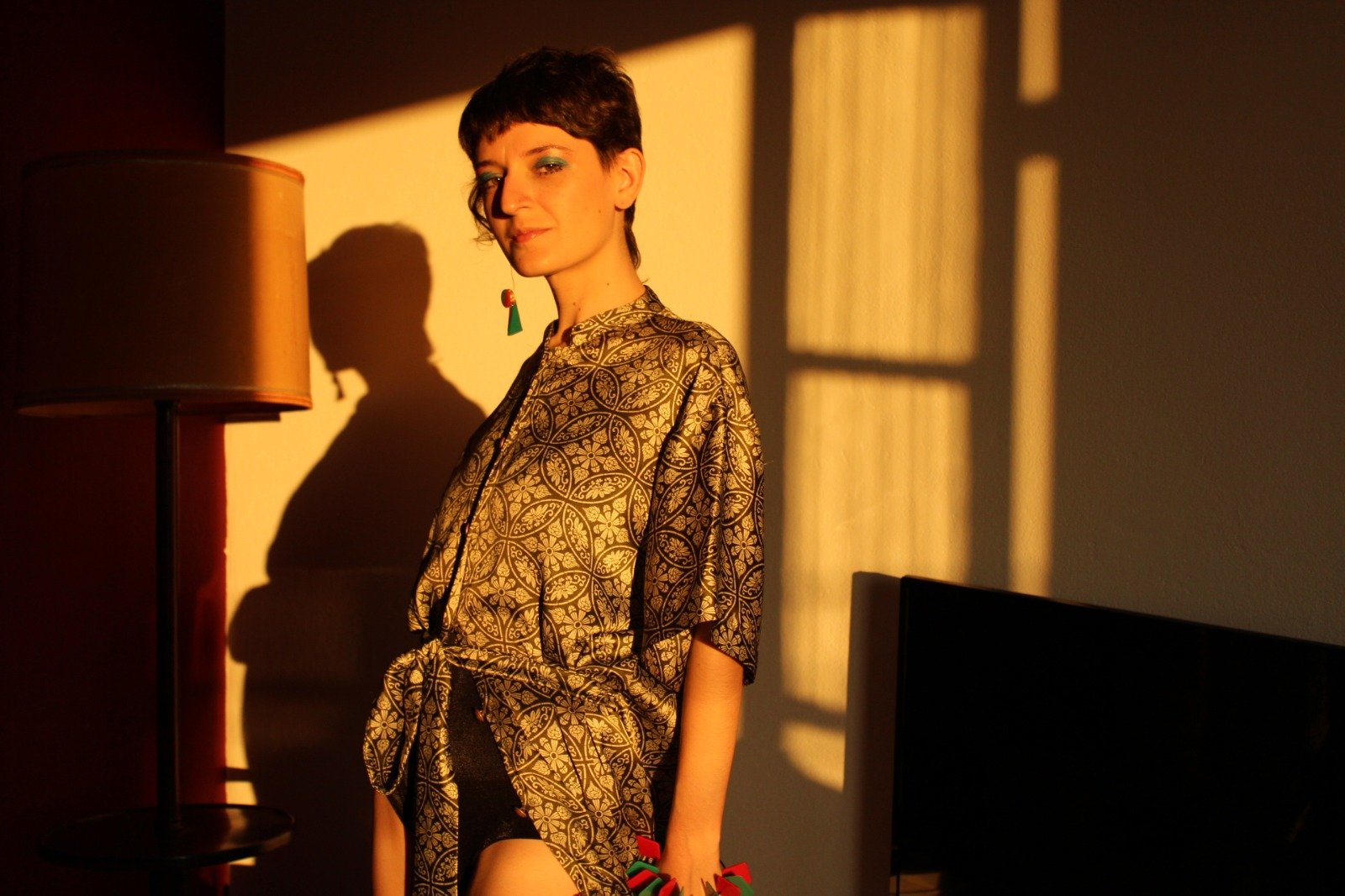 Mulher posando em uma sala iluminada pelo sol, vestindo uma túnica com estampa dourada e brincos coloridos. Ao fundo, há uma sombra de luminária e janela projetada na parede.