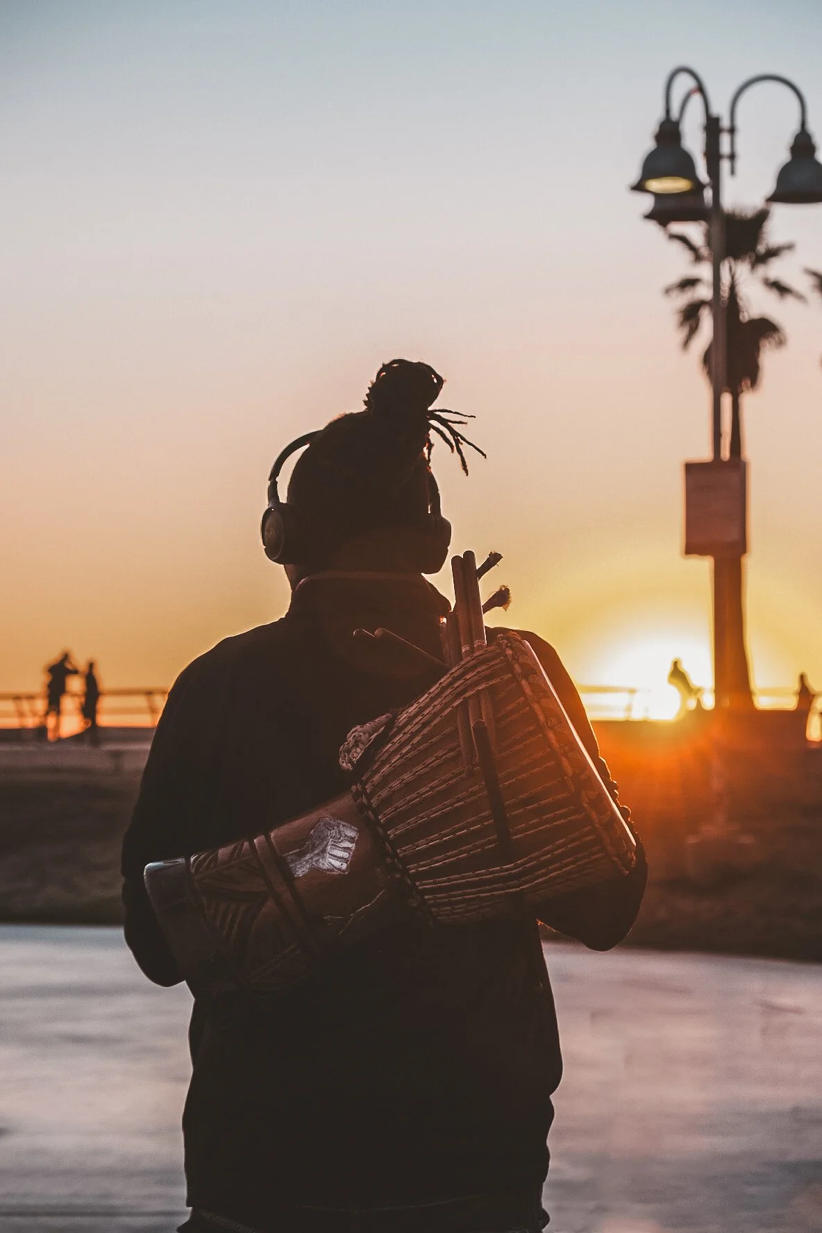 Person wearing headphones with a drum on their back, standing near a beach at sunset at venice beach california.