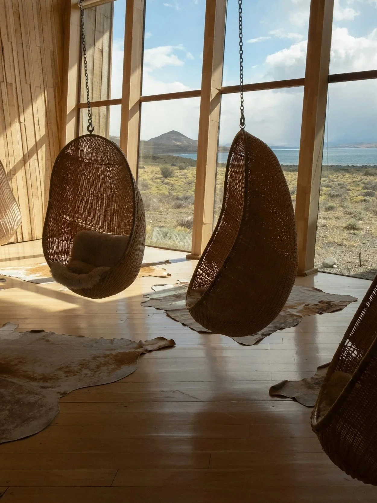 Two hanging wicker egg chairs inside a wooden room with large windows showing a desert landscape and mountains in the background.