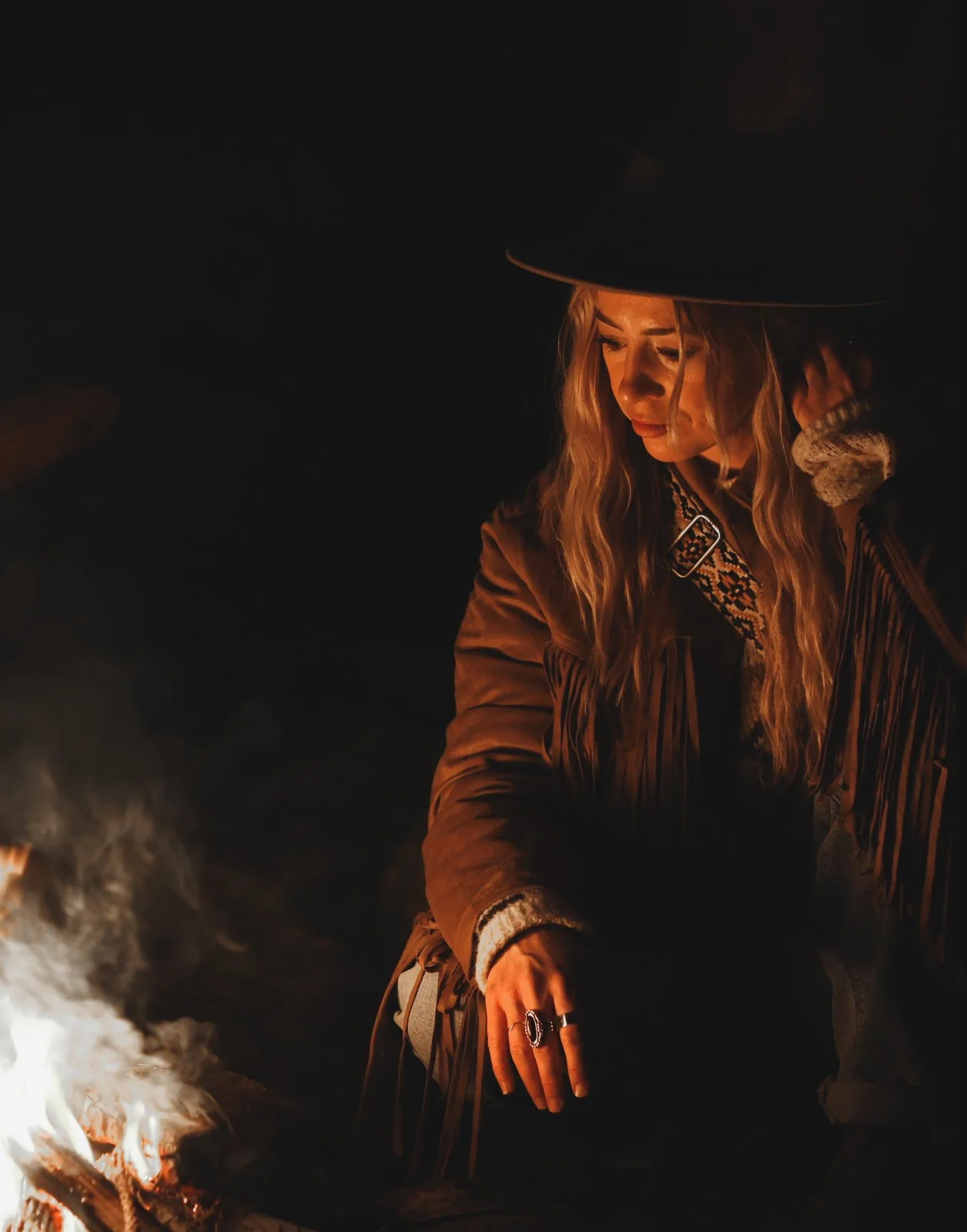 Woman with long blonde hair and a hat sitting by a campfire at night