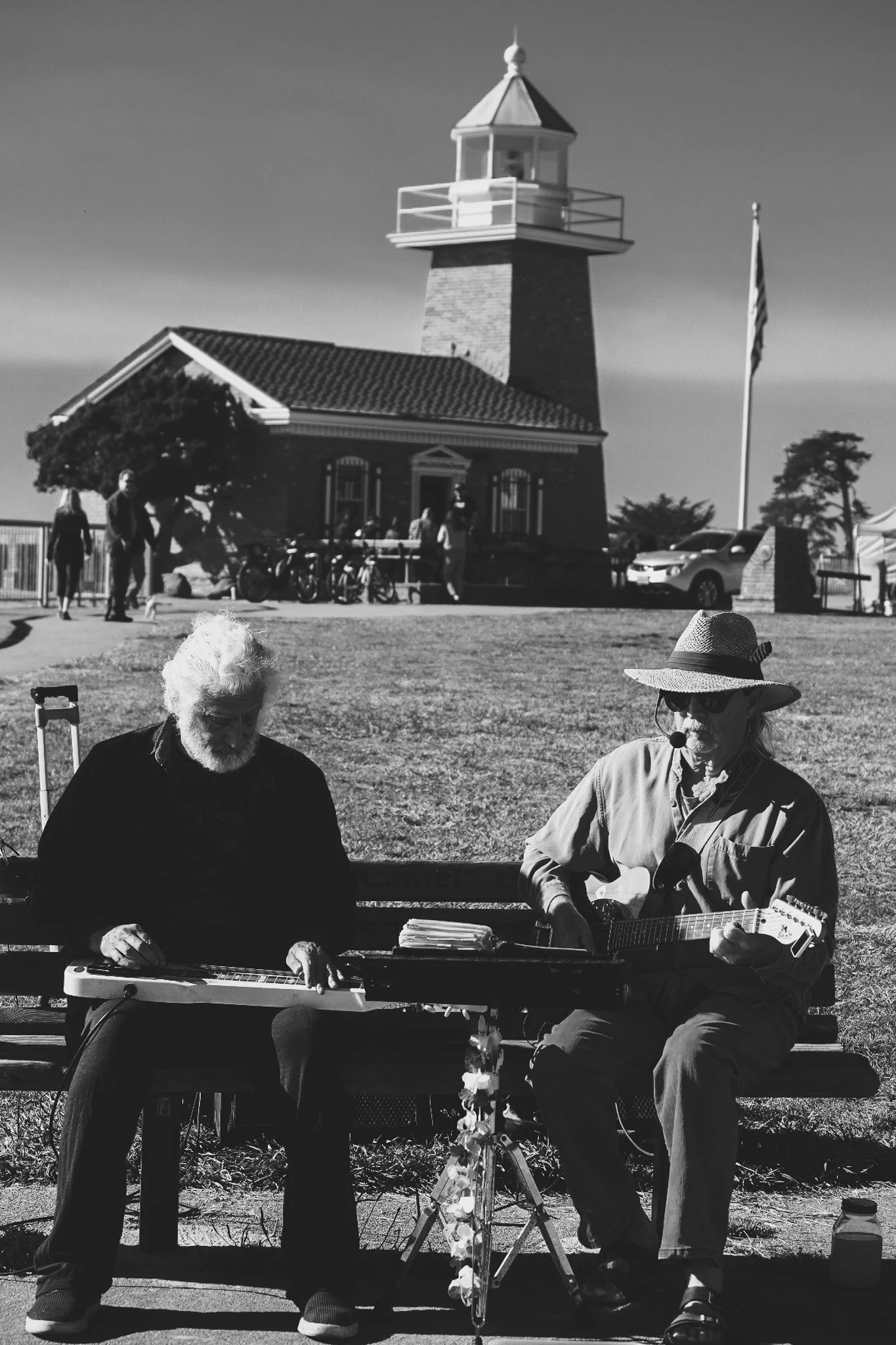 Two elderly men playing instruments on a bench in front of a lighthouse, one playing a slide guitar and the other an electric guitar, with American flag and building in background, in black and white in santa cruz