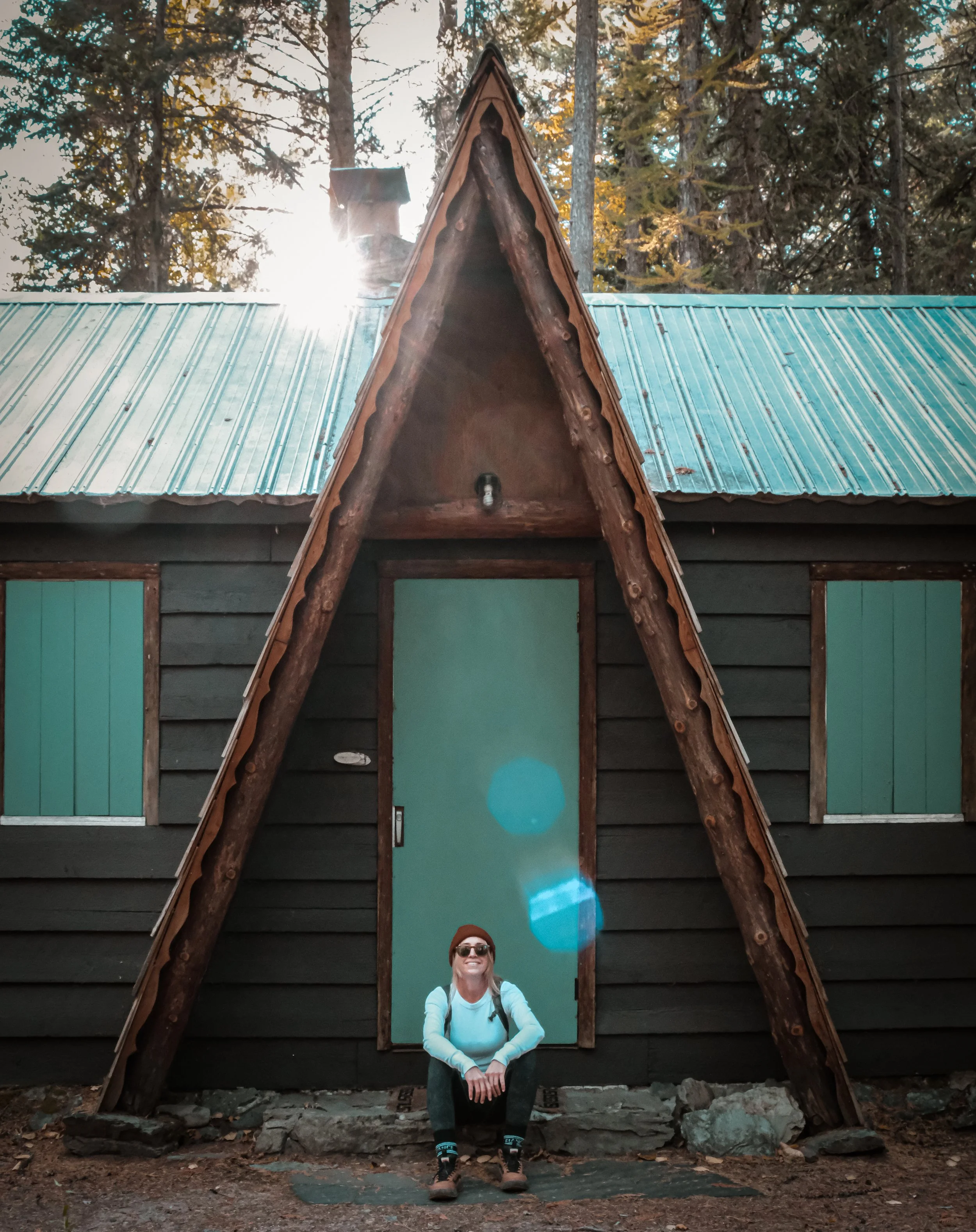 A woman sits smiling in front of an A-frame cabin with dark wooden siding and green window shutters, surrounded by tall pine trees in a forest.