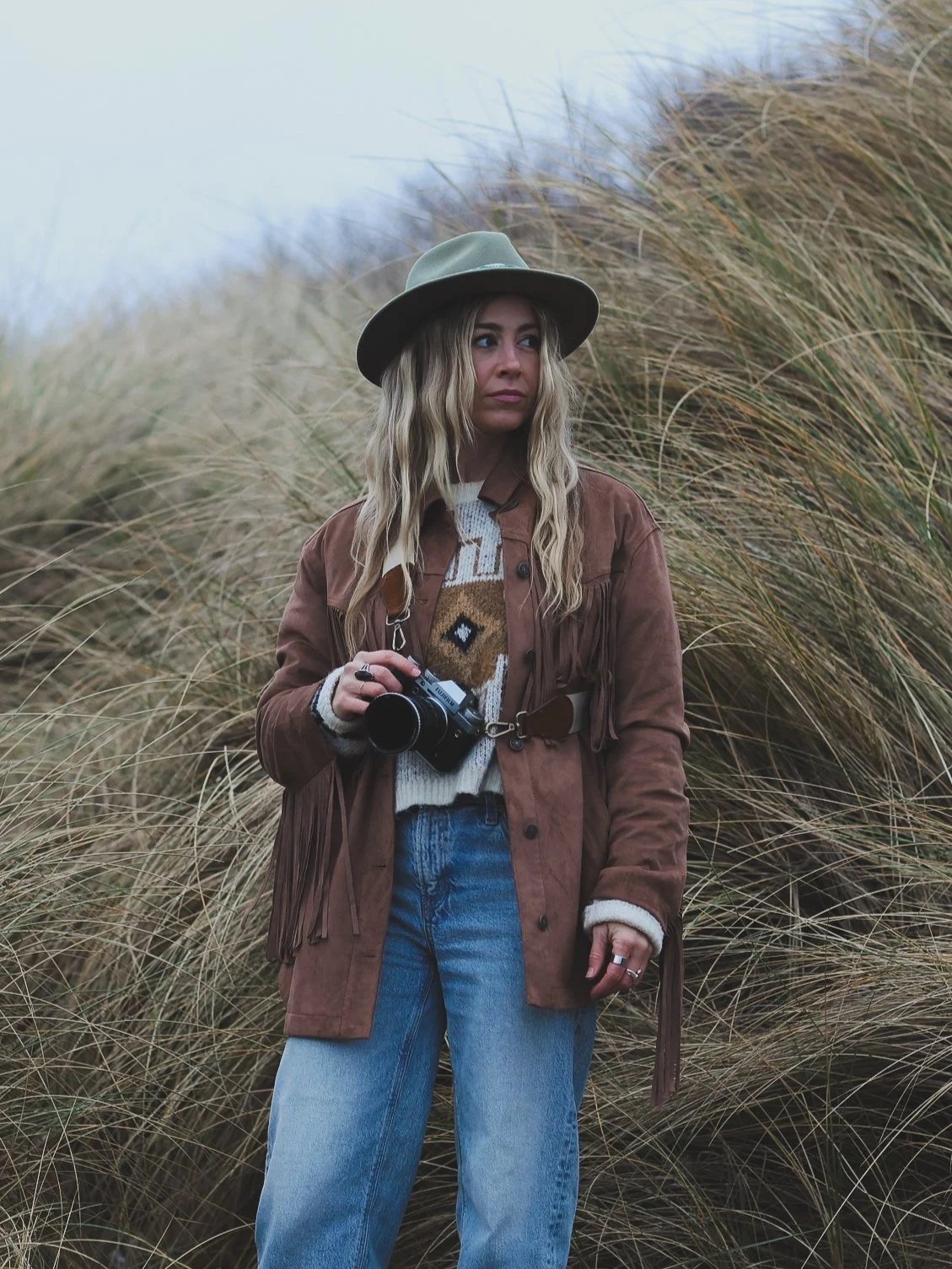 A woman standing in front of tall grass, wearing a hat, a brown fringed jacket, a patterned sweater, and blue jeans, holding a camera.