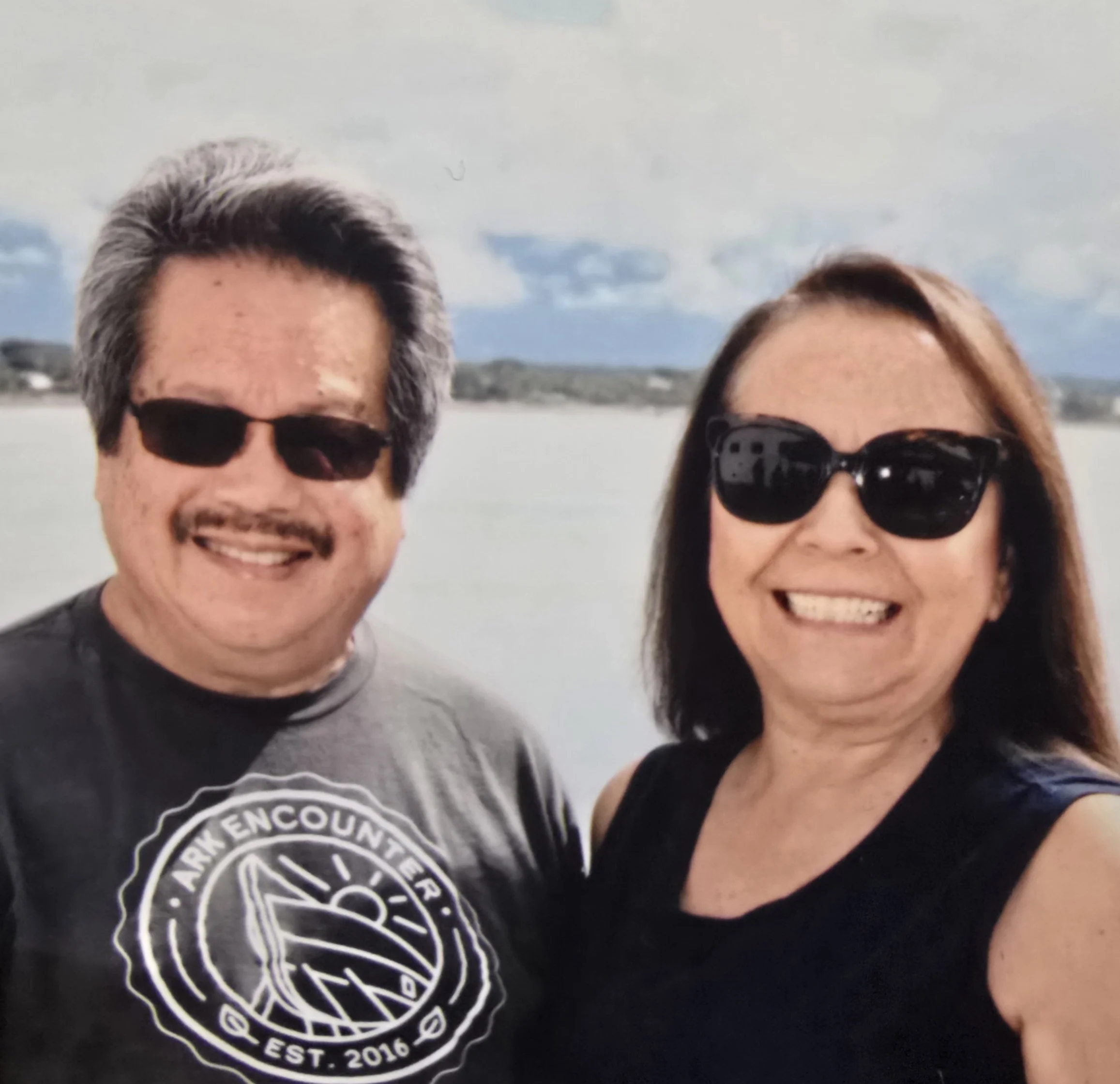 A smiling man and woman wearing sunglasses taking a selfie outdoors with water and land in the background.