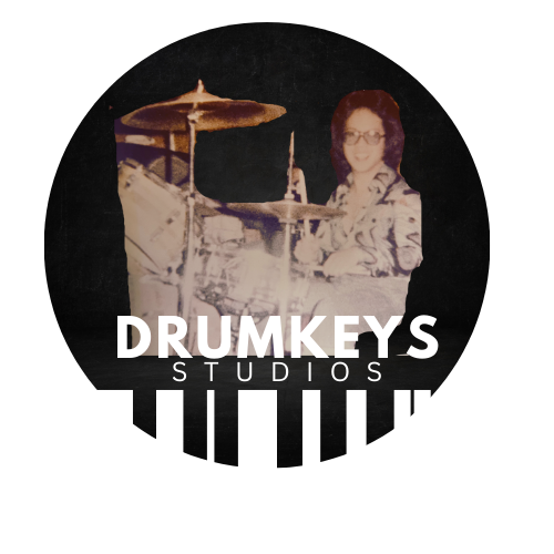 Logo for Drumkeys Studios featuring a woman with curly hair and glasses sitting behind a drum set, with the studio name in bold white letters over a black circular background.