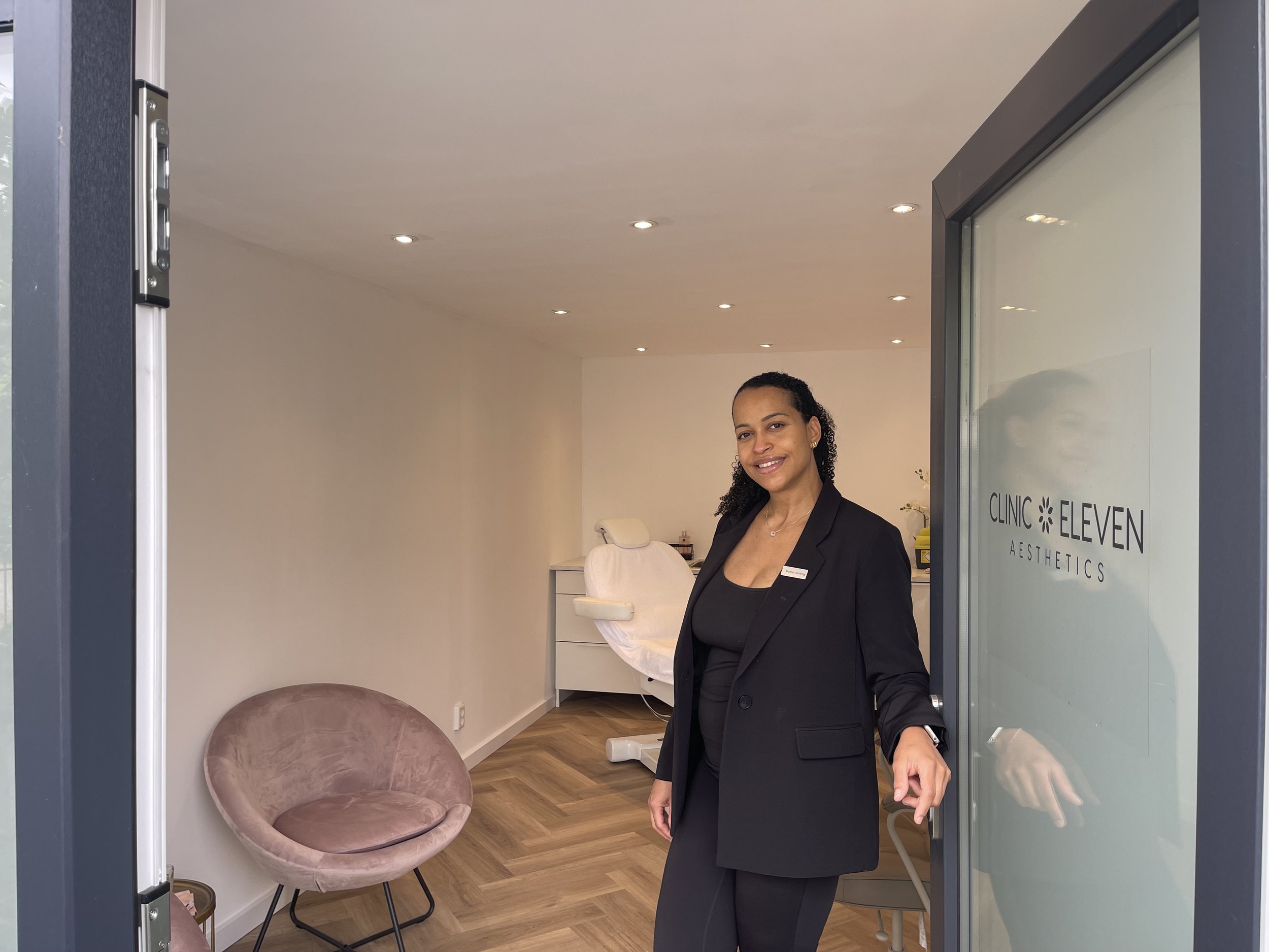 A woman in a black blazer and pants stands at the doorway of a clinic, smiling. The clinic's sign reads 'CLINIC ELEVEN AESTHETICS.' Inside, there is a pink chair and a treatment bed with a white cover in a clean, minimal room with wood flooring and recessed lighting.
