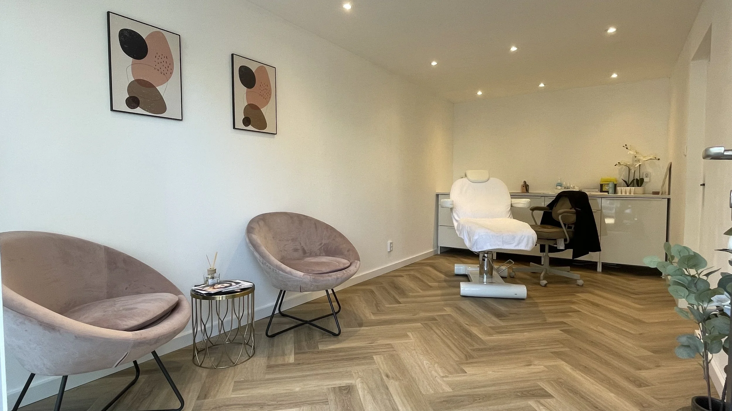 A clean, modern medical or dental office waiting room with two beige velvet chairs, a round metal side table with magazines, a white examination chair, and a small countertop with medical supplies and plants in the background.
