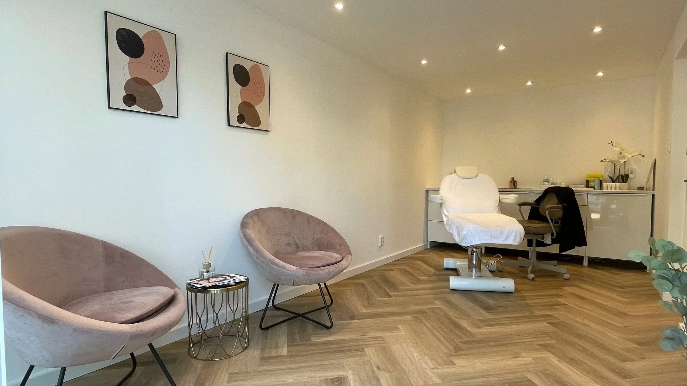 A modern, clean room with two pastel pink velvet chairs, a small round golden side table, and a white medical examination chair with a headrest. Wall art with abstract shapes hangs on the white wall, and ceiling lights are visible. There is a desk in the background with various items, including a potted plant and office supplies.