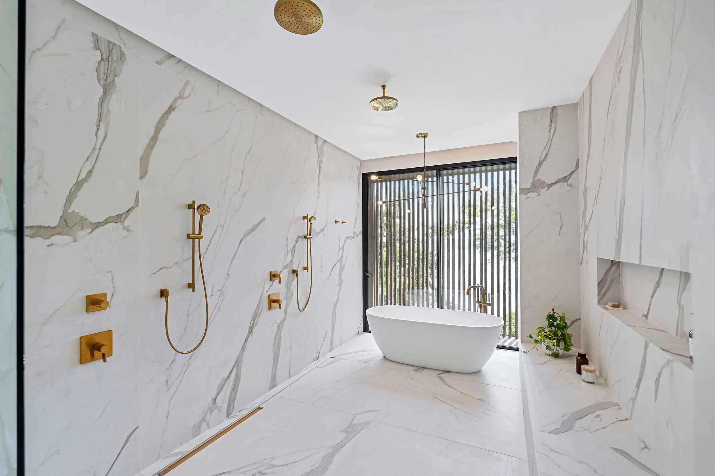 Modern luxury bathroom with marble walls, freestanding bathtub, gold fittings, and large window with vertical blinds.