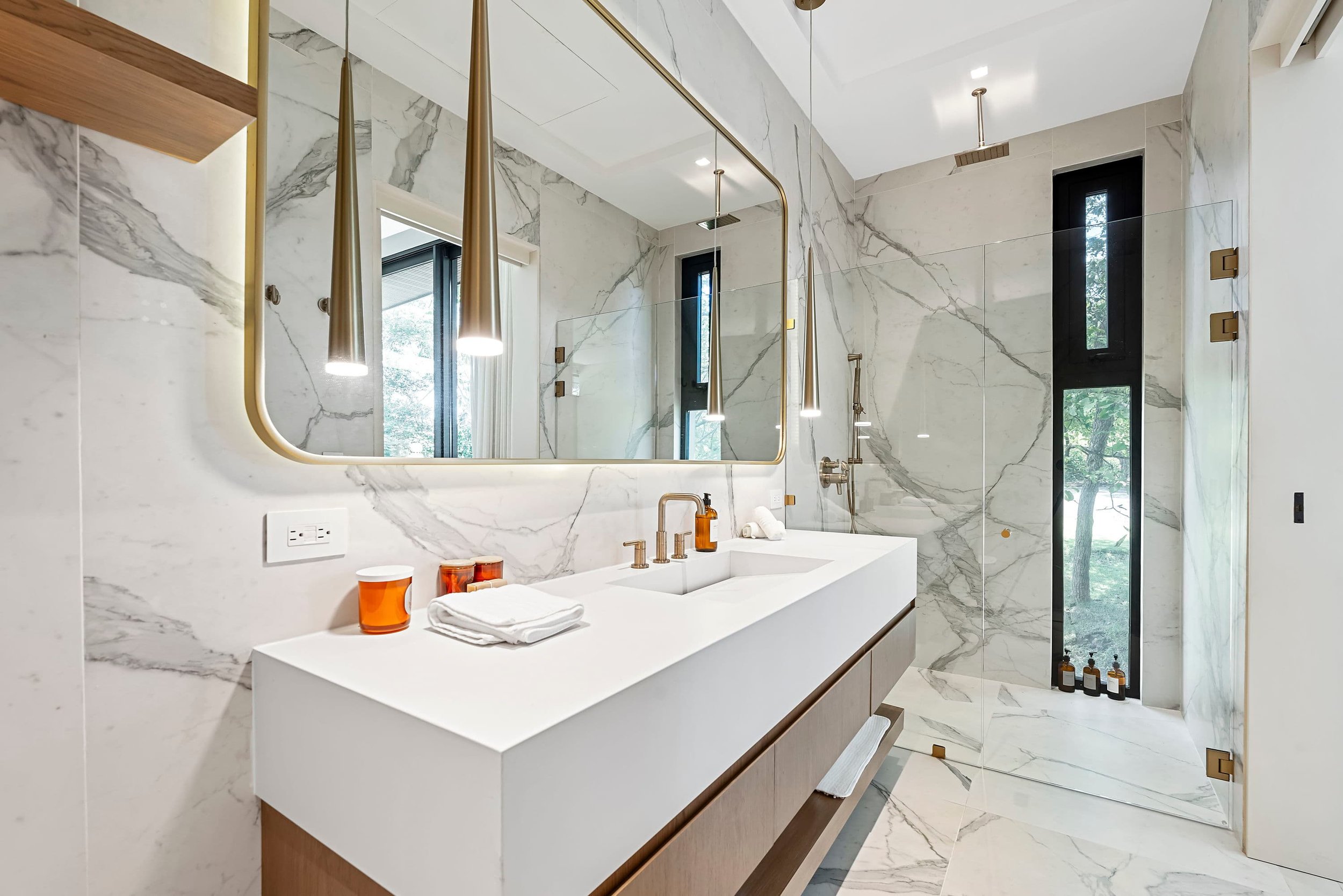 Modern bathroom with marble walls, a large mirror, and floating vanity. It features pendant lights, a white countertop, gold fixtures, and a walk-in glass shower.
