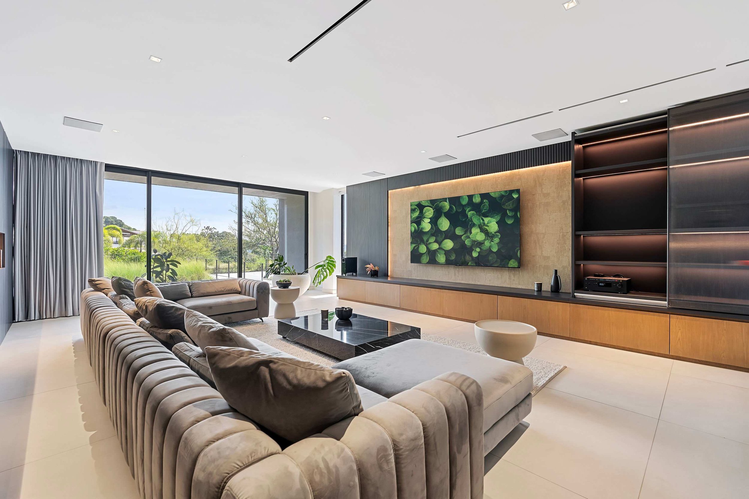 Modern living room with large sectional sofa, glass coffee table, potted plants, wall-mounted flat-screen TV, and floor-to-ceiling windows overlooking greenery.