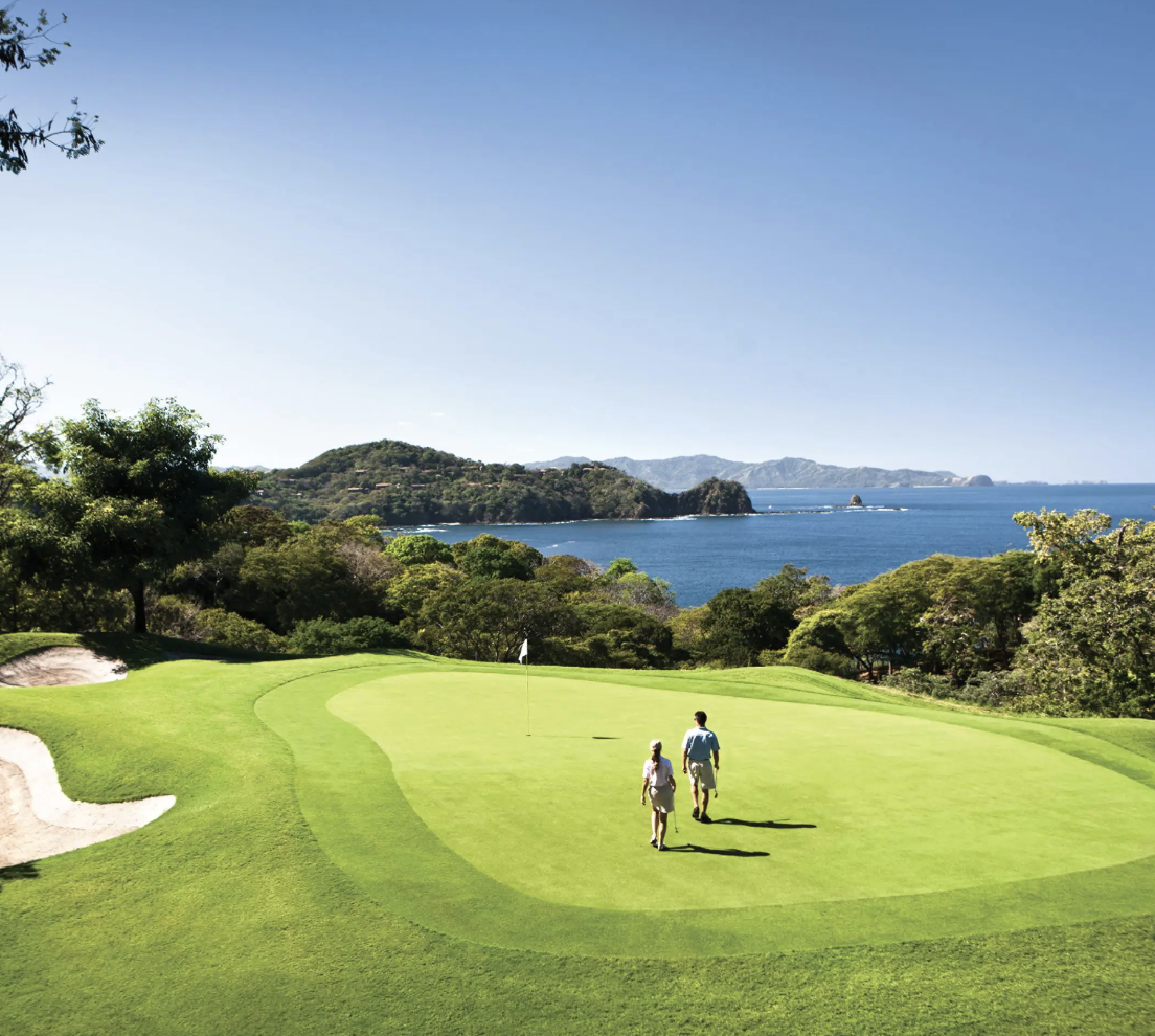 Two people walking on a golf course near the ocean with a view of the coastline and hills in the background at Four Seasons Papagayo.